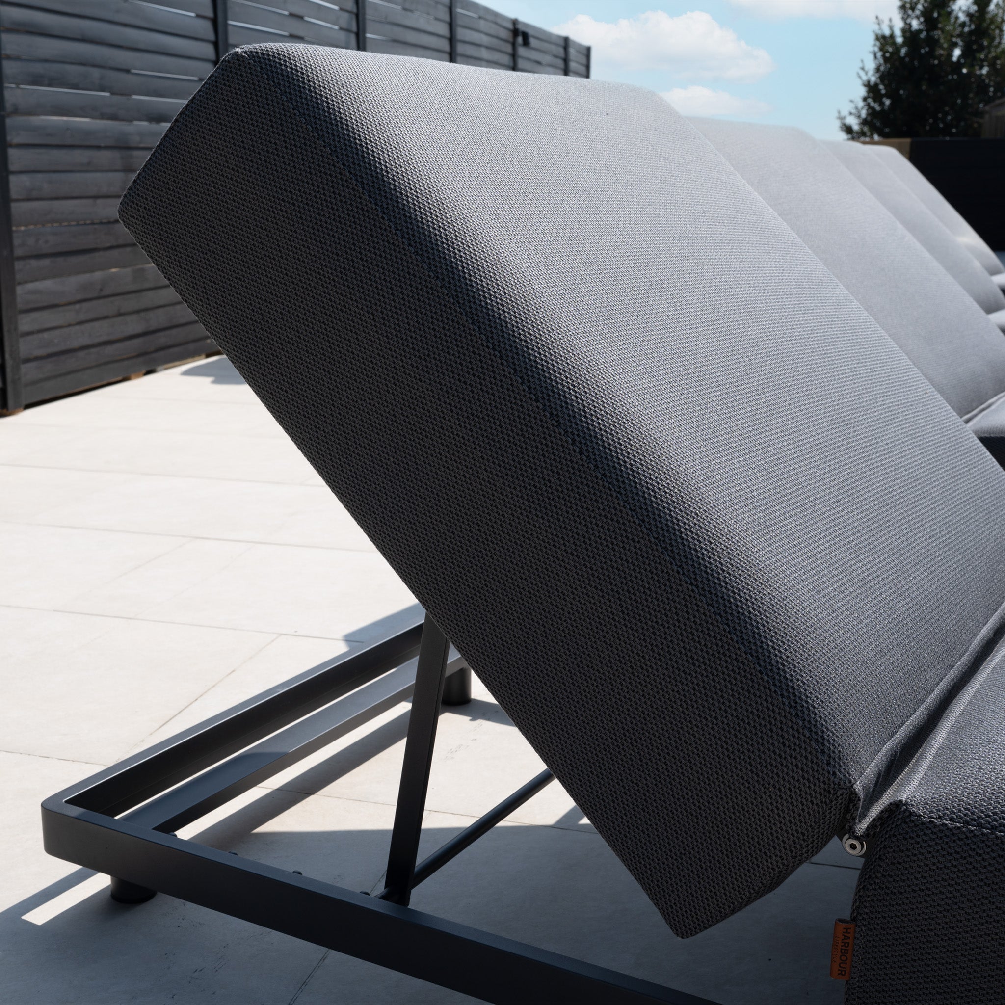 Close-up of modern grey Luna Outdoor Fabric Deluxe Sun Lounger with Bolster Cushion on a sunny patio, featuring durable solution-dyed acrylic fabric and sleek black frames for lasting outdoor comfort.