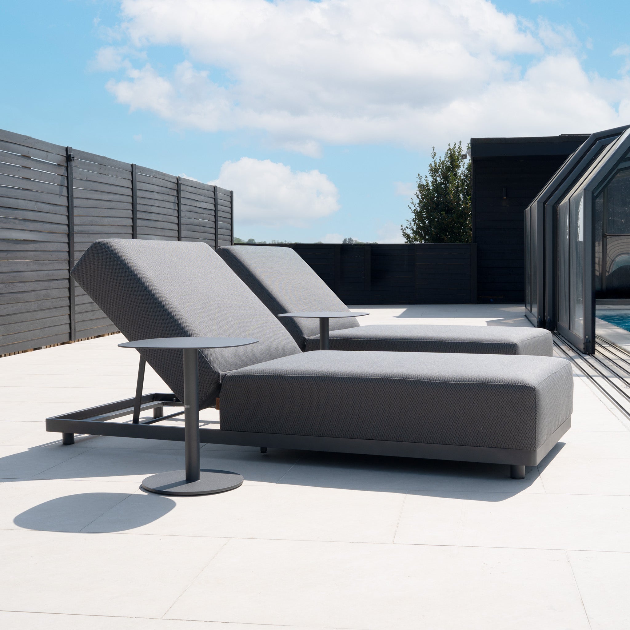 A Luna Outdoor Fabric Deluxe Sun Lounger with Bolster Cushion in Grey sits on a spacious patio with a black fence and blue sky, paired with a small table.