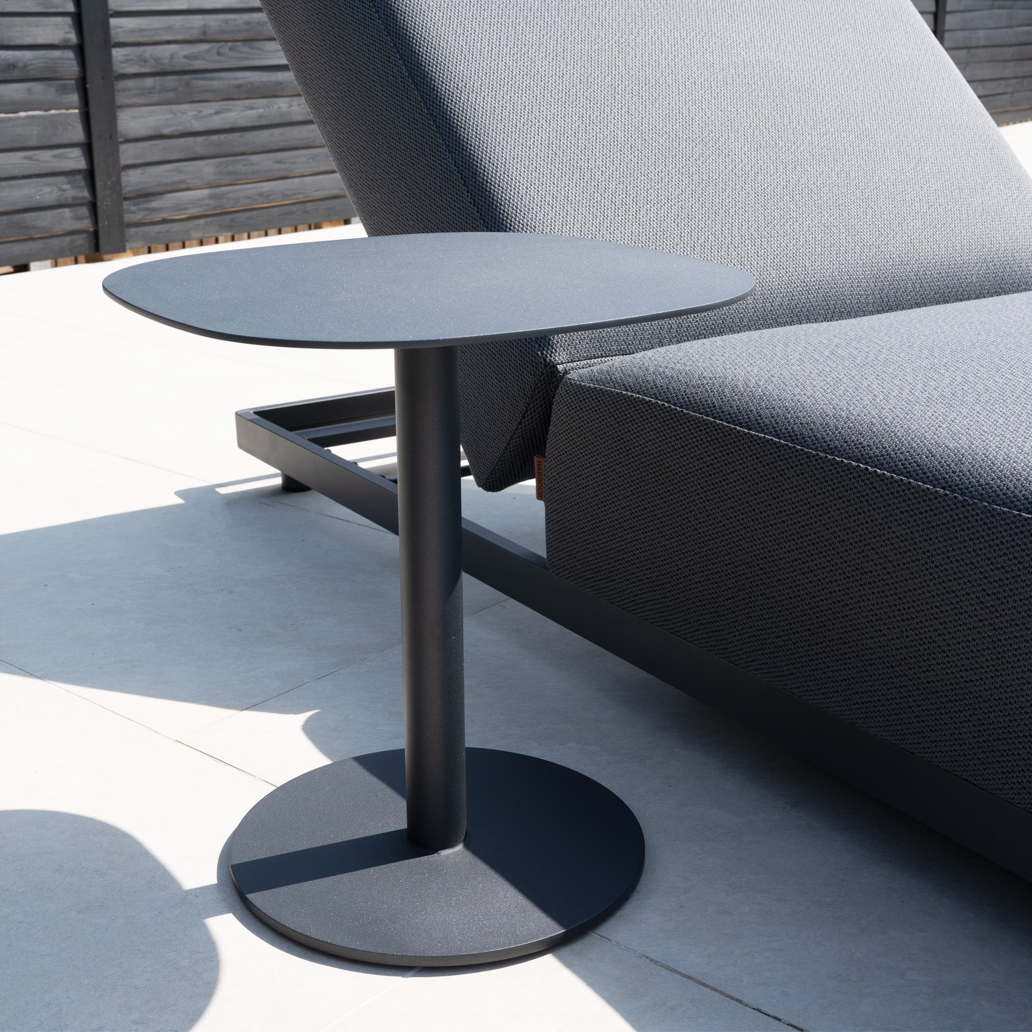 A round black side table sits beside the Luna Outdoor Fabric Deluxe Sun Lounger with Bolster Cushion in Grey on a tiled patio.