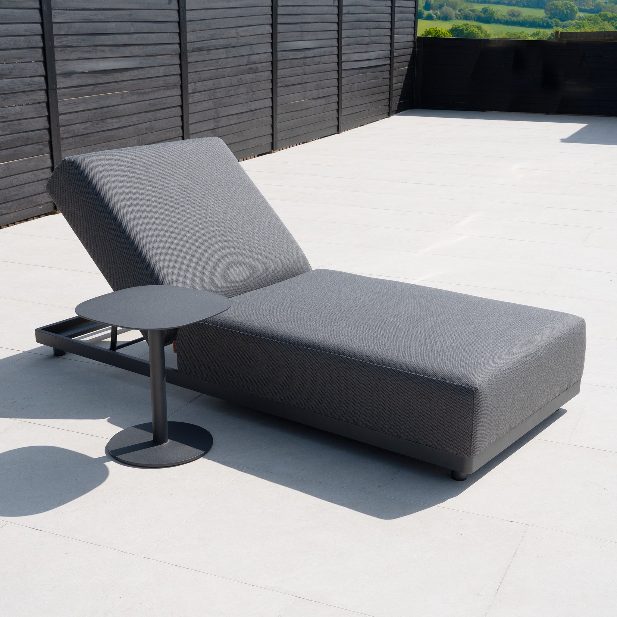 A Luna Outdoor Fabric Deluxe Sun Lounger with Bolster Cushion in Grey sits beside a small round side table on a spacious patio with black fencing.