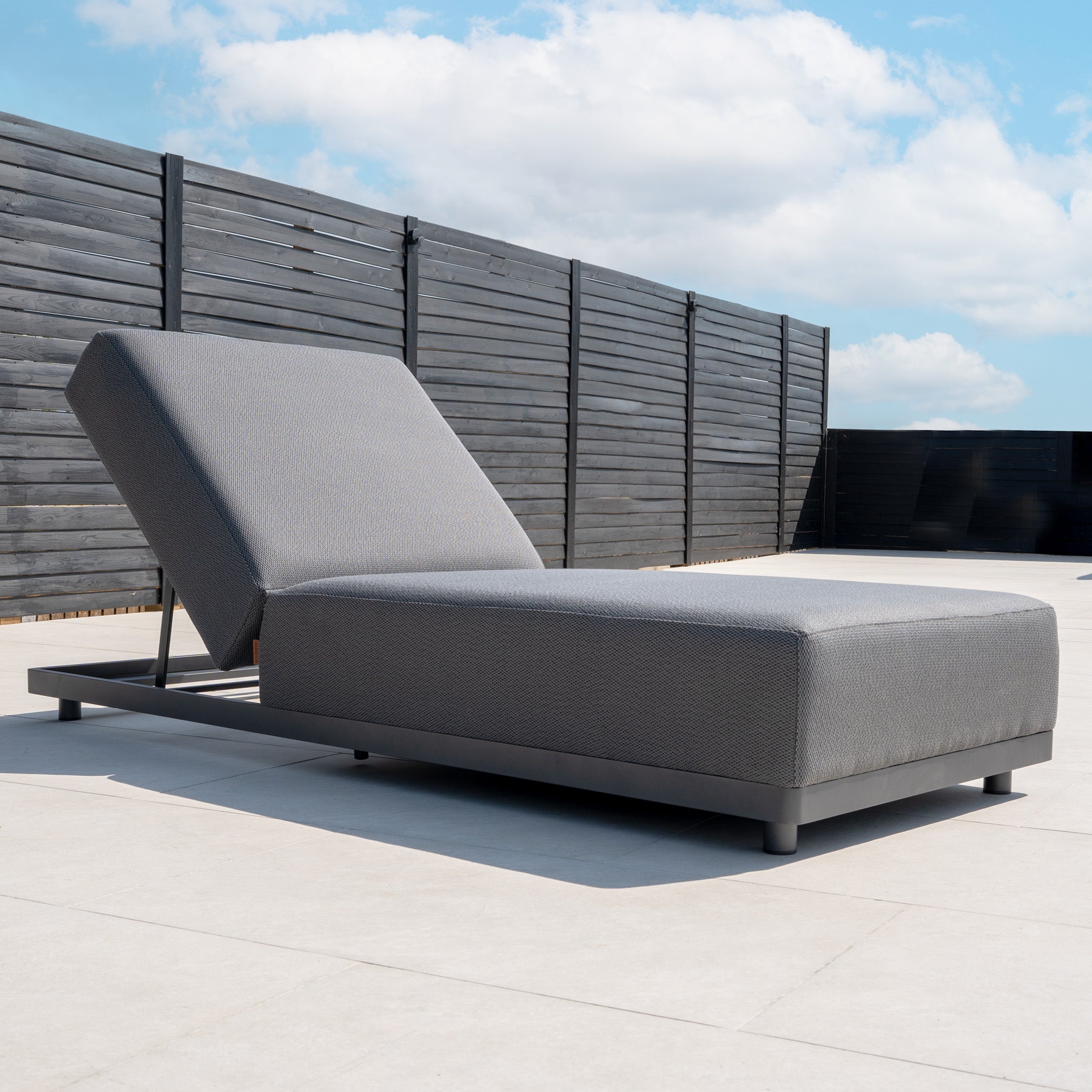 The Luna Outdoor Fabric Deluxe Sun Lounger with Bolster Cushion in Grey is featured on a spacious patio, set against a black privacy fence and a blue sky with clouds.