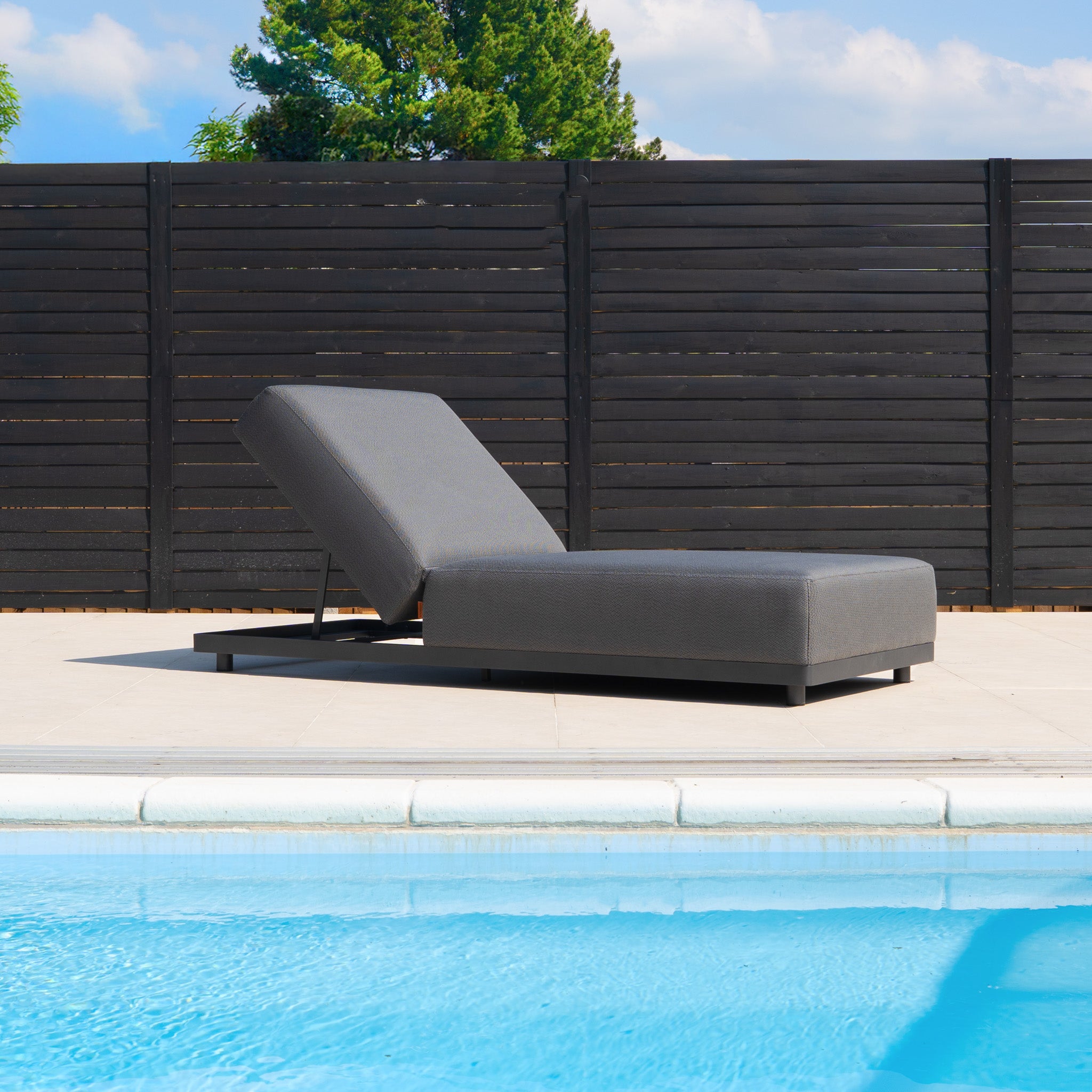 The Luna Outdoor Fabric Deluxe Sun Lounger with Bolster Cushion in Grey sits on a patio by a black wooden fence and trees, featuring stain-resistant outdoor fabric for lasting durability.