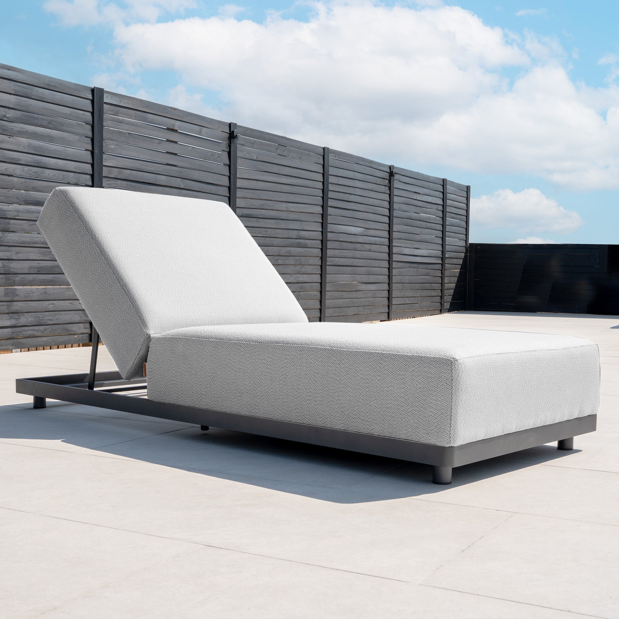The Luna Outdoor Fabric Deluxe Sun Lounger with Bolster Cushion in Oyster Grey, featuring an adjustable backrest, is placed on a tiled patio near a black fence beneath a blue sky with clouds.