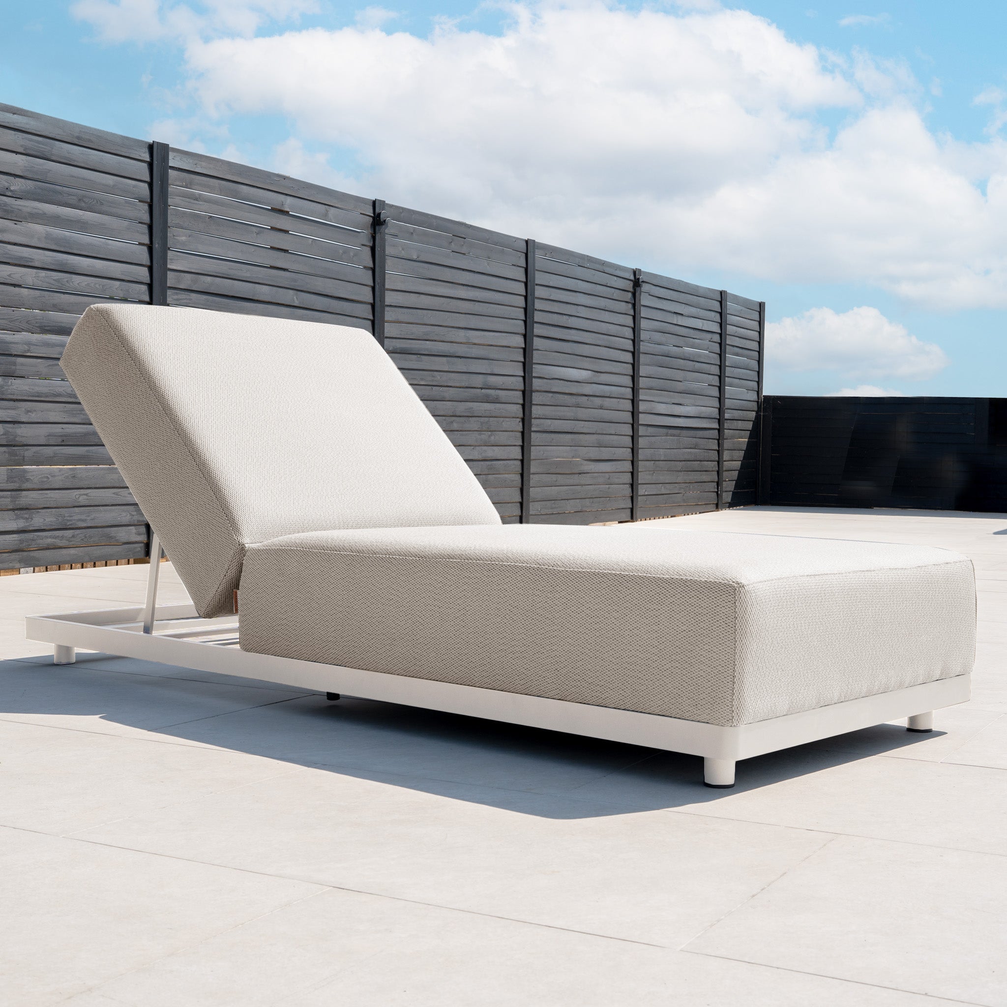 The Luna Outdoor Fabric Deluxe Sun Lounger with Bolster Cushion in Fawn rests on a sunlit patio, framed by a dark wooden fence and blue sky, crafted from weatherproof, stain-resistant materials for lasting outdoor comfort.