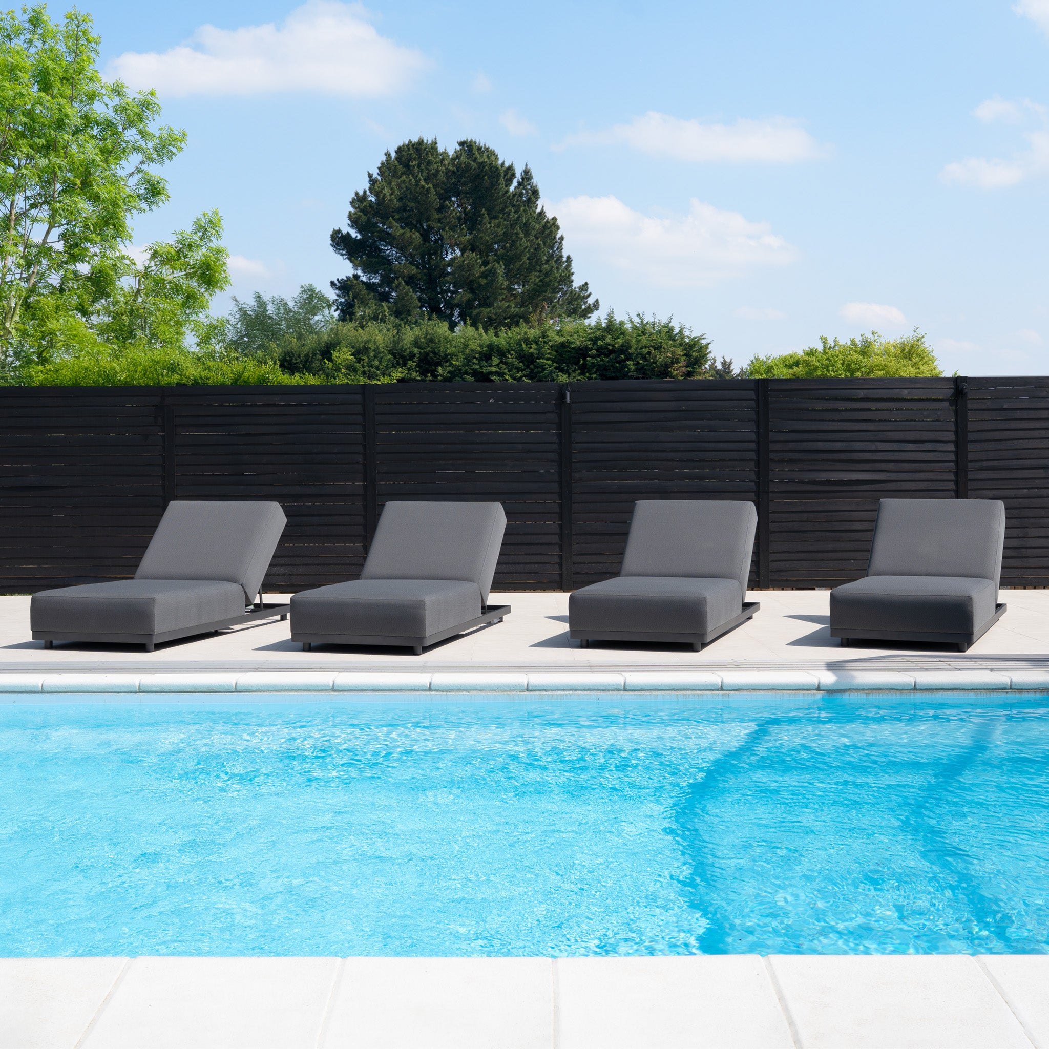 Four Luna Outdoor Fabric Deluxe Sun Lounger with Bolster Cushion in Grey line a clear blue pool, surrounded by trees and a sleek black fence finished with TIGER Drylac® Powder Coating.