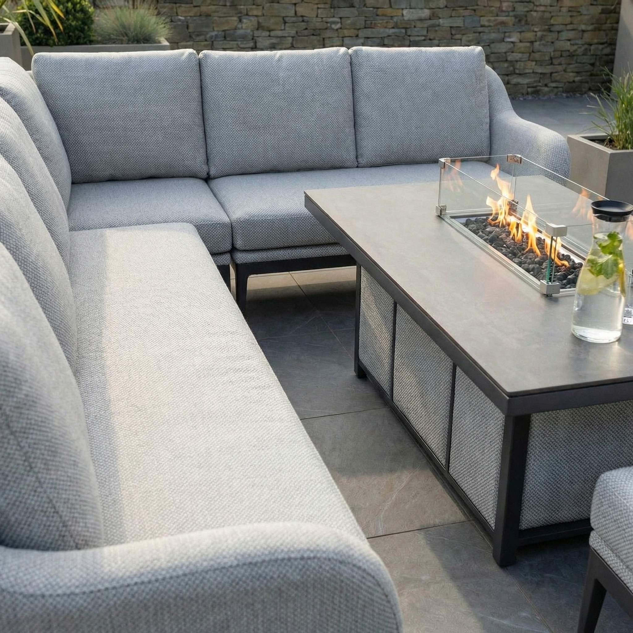 The Luna Outdoor Fabric Rectangular Corner Dining Set with Rising Firepit Table in Oyster Grey (Right Hand) is shown on a stone patio, with a drink placed on the table.
