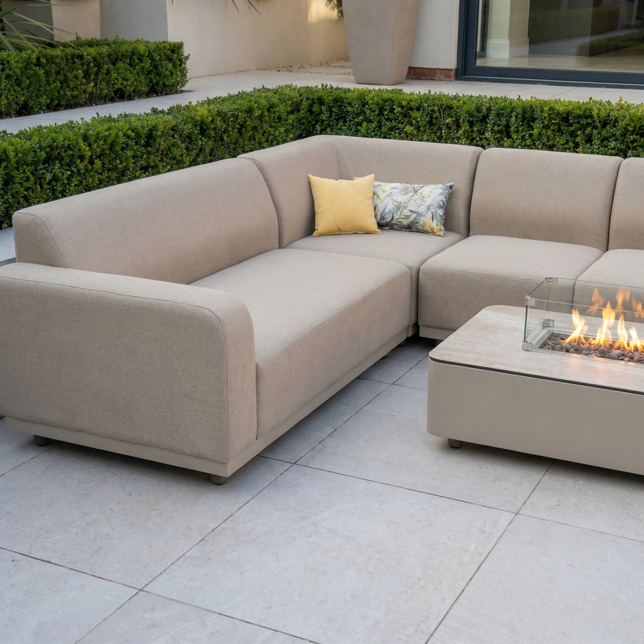 Luna Outdoor Fabric Large Corner Group Set with Firepit Coffee Table in Fawn arranged on a tiled patio with greenery in the background.