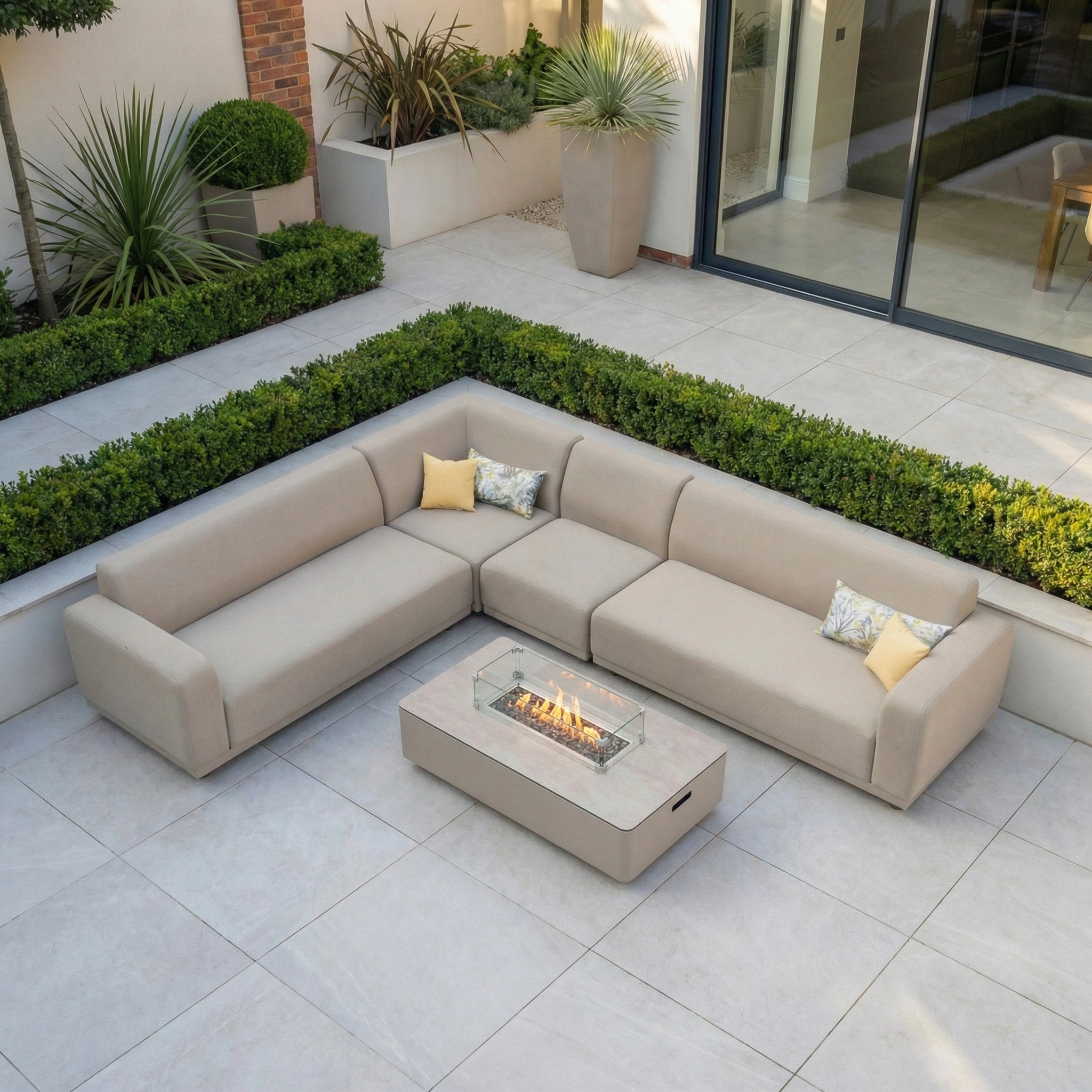The Luna Outdoor Fabric Large Corner Group Set with Firepit Coffee Table in Fawn is shown surrounded by greenery on a tiled patio.