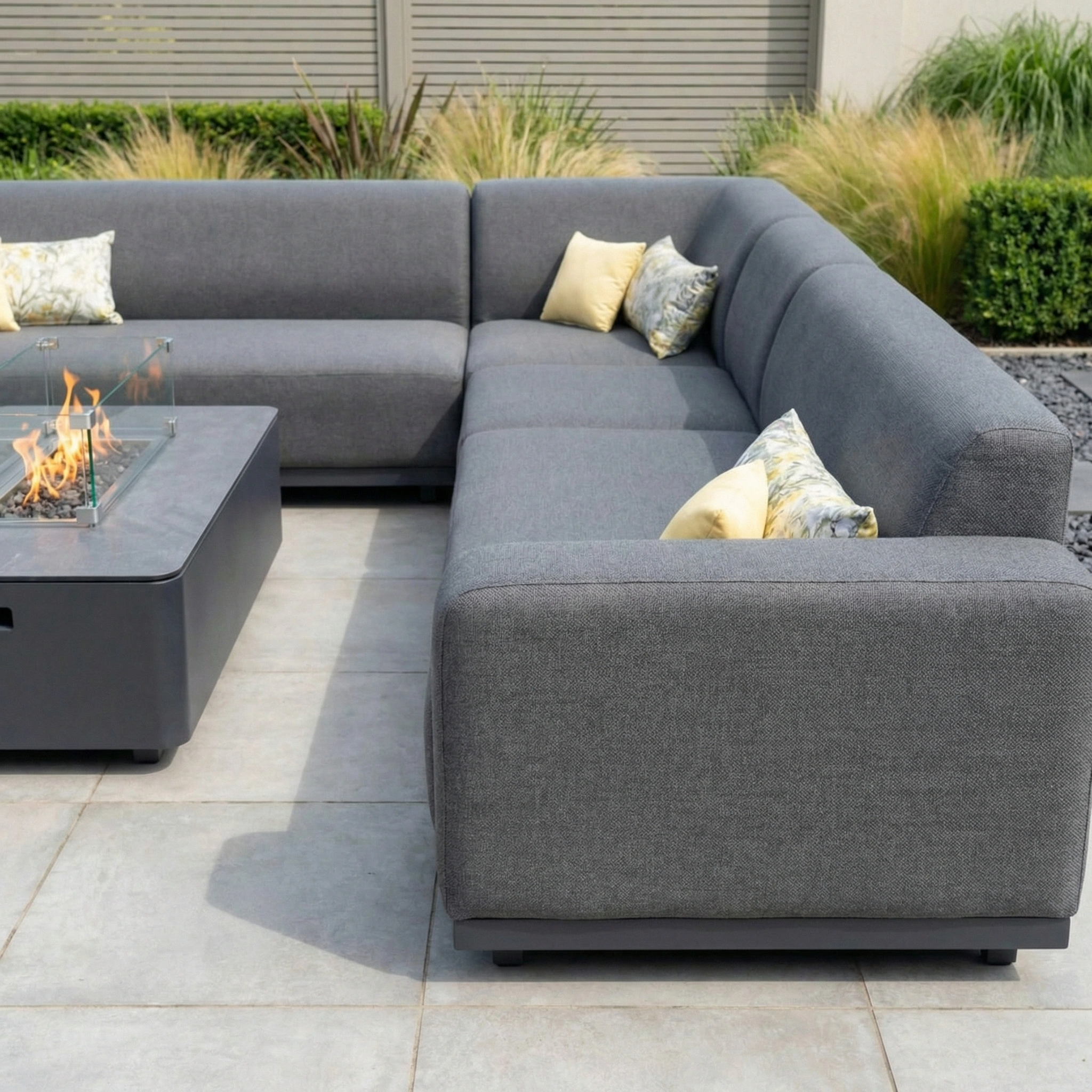 The Luna Outdoor Fabric Large Corner Group Set with Firepit Coffee Table in Grey creates a cozy outdoor retreat, featuring stylish seating arranged around the firepit on a tiled patio.