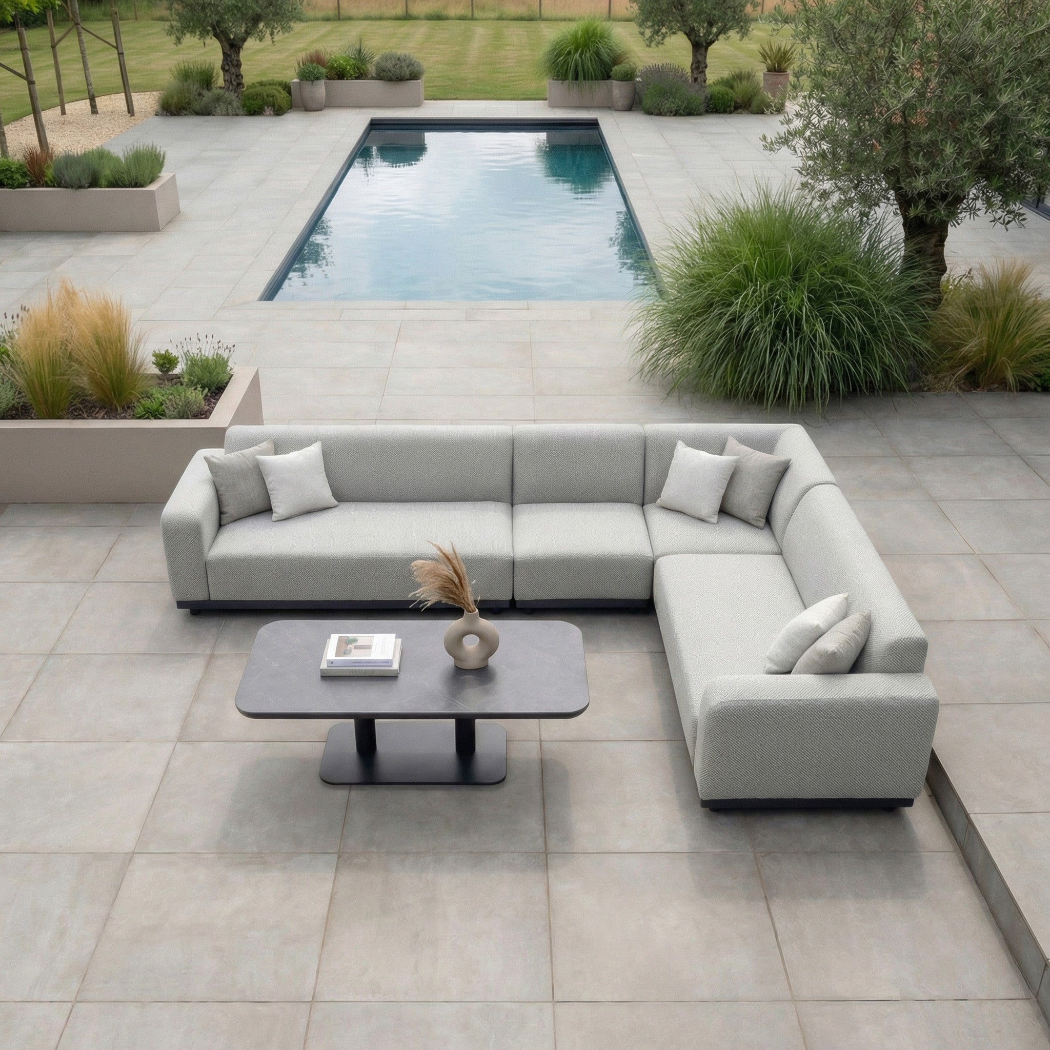 Modern outdoor patio featuring the Luna Outdoor Fabric Large Corner Sofa Set with Altura Rising Table in Oyster Grey, plus a pool surrounded by greenery and planters.