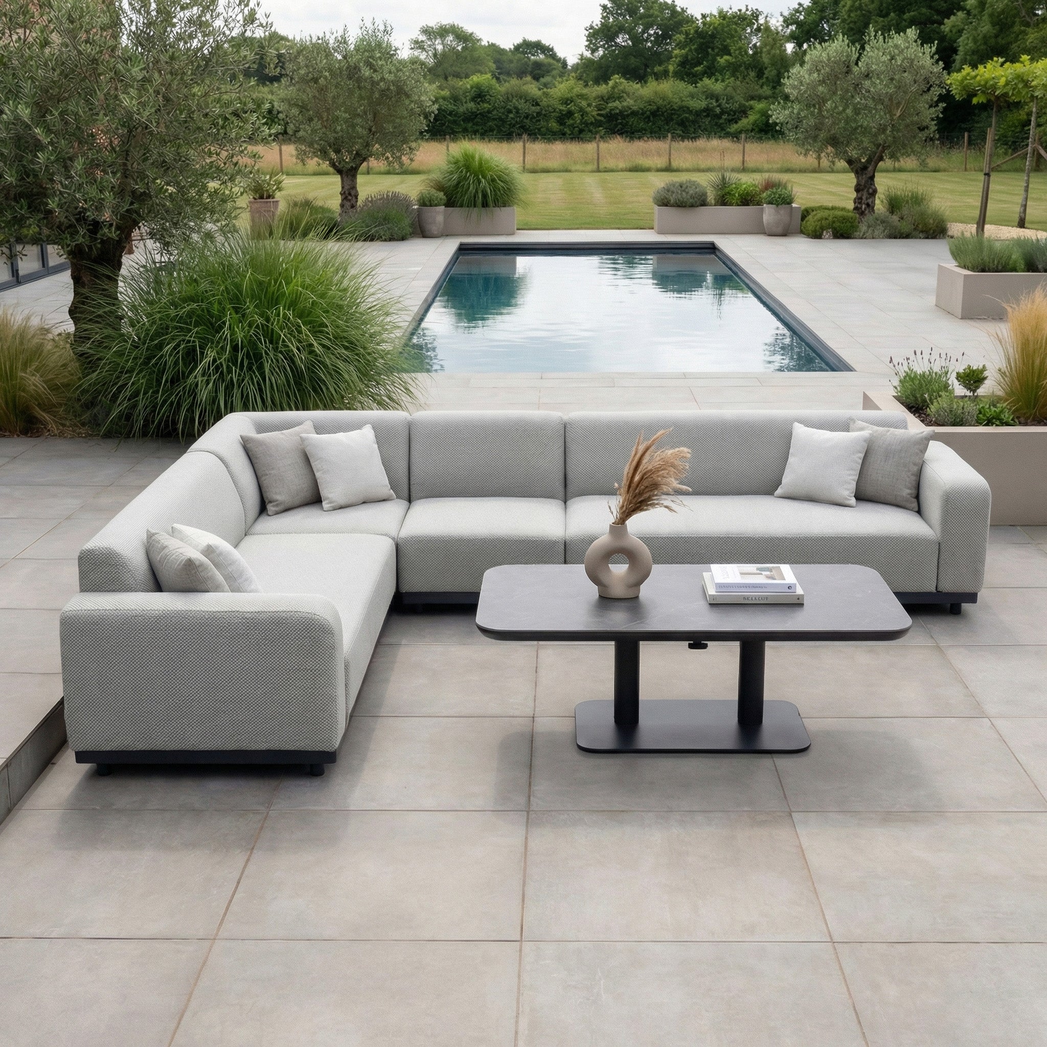 A modern patio with the Luna Outdoor Fabric Large Corner Sofa Set and Altura Rising Table in Oyster Grey, plus a pool surrounded by greenery and planters.