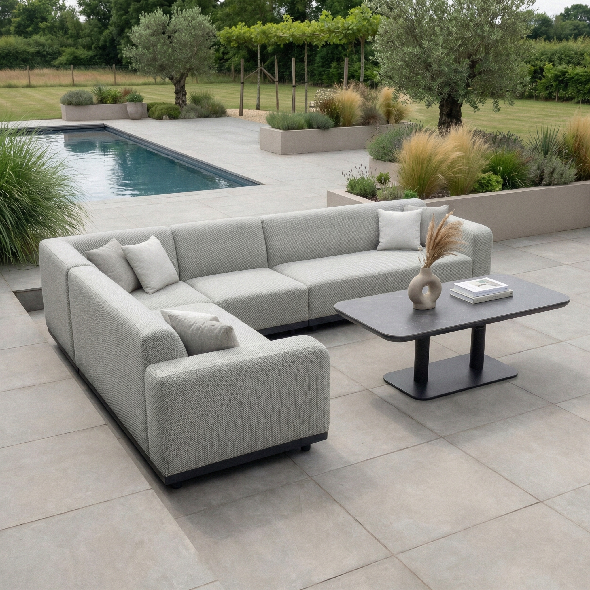 Modern patio with a Luna Outdoor Fabric Large Corner Sofa Set in Oyster Grey and matching Altura Rising Table, set beside a pool and surrounded by lush greenery and planters.