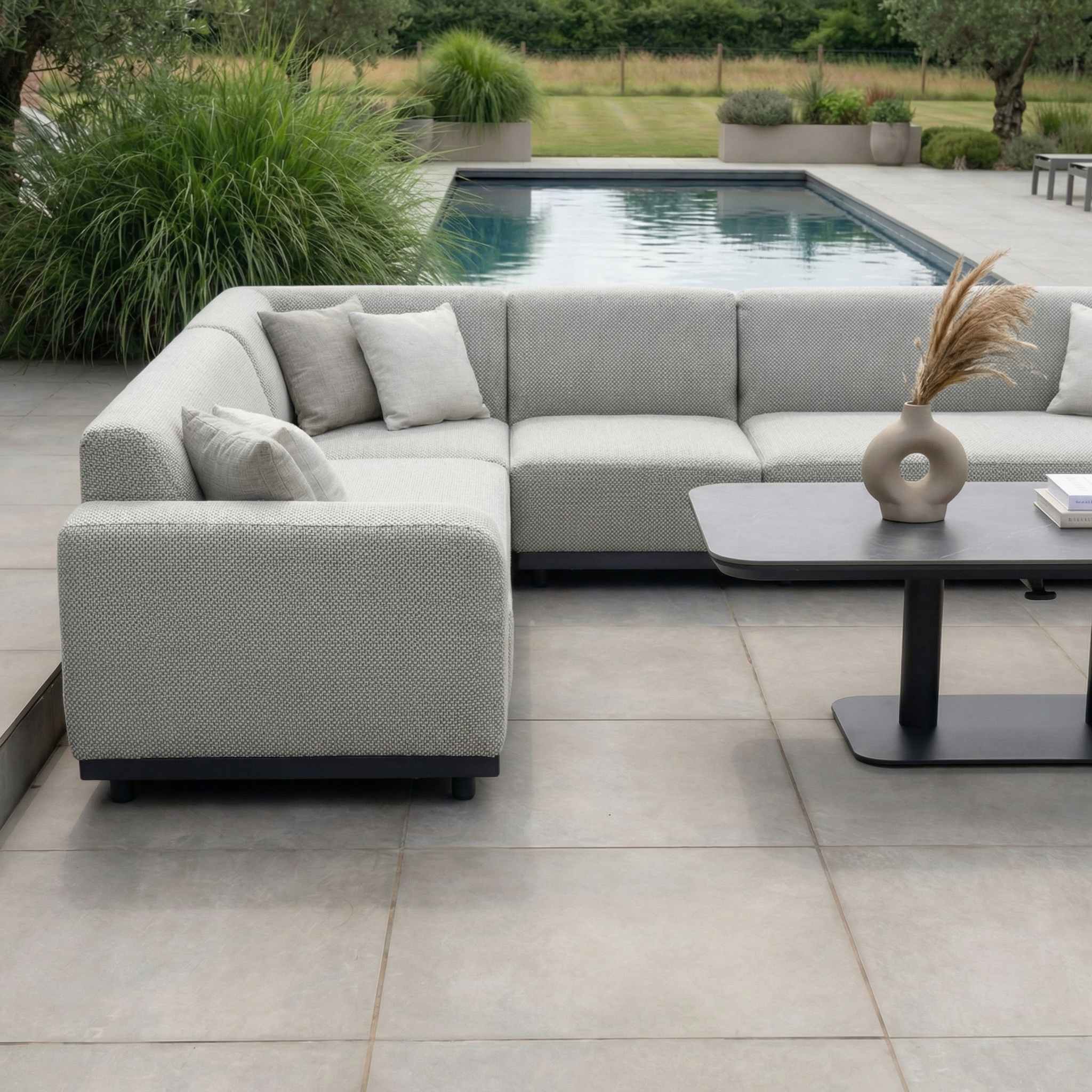 Contemporary outdoor seating area featuring the Luna Outdoor Fabric Large Corner Sofa Set with Altura Rising Table in Oyster Grey, plus a pool bordered by greenery.