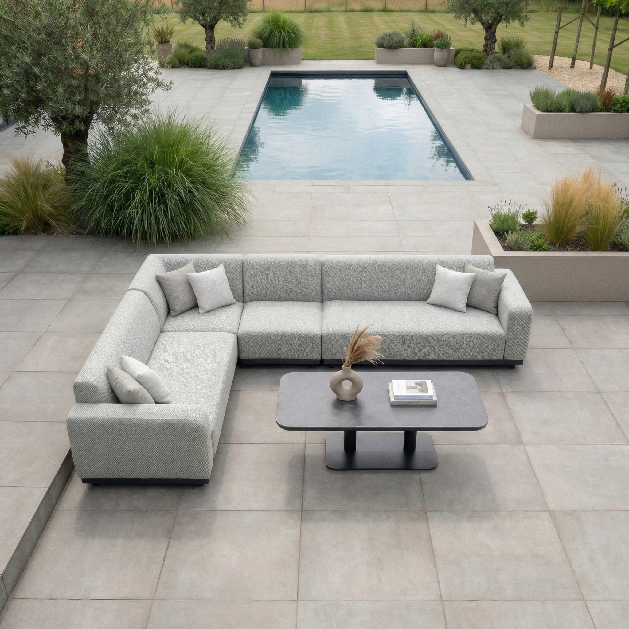 Modern patio featuring the Luna Outdoor Fabric Large Corner Sofa Set with Altura Rising Table in Oyster Grey, paired with a coffee table and a pool surrounded by greenery.