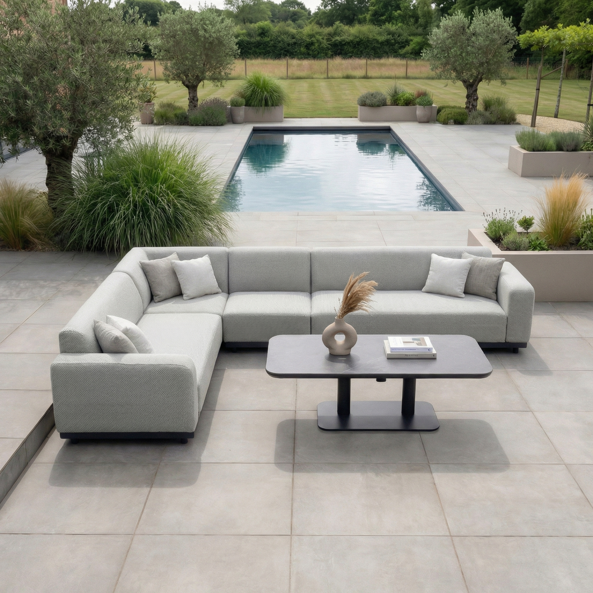 Modern outdoor patio featuring the Luna Outdoor Fabric Large Corner Sofa Set with Altura Rising Table in Oyster Grey, plus a pool bordered by lush greenery and trees.