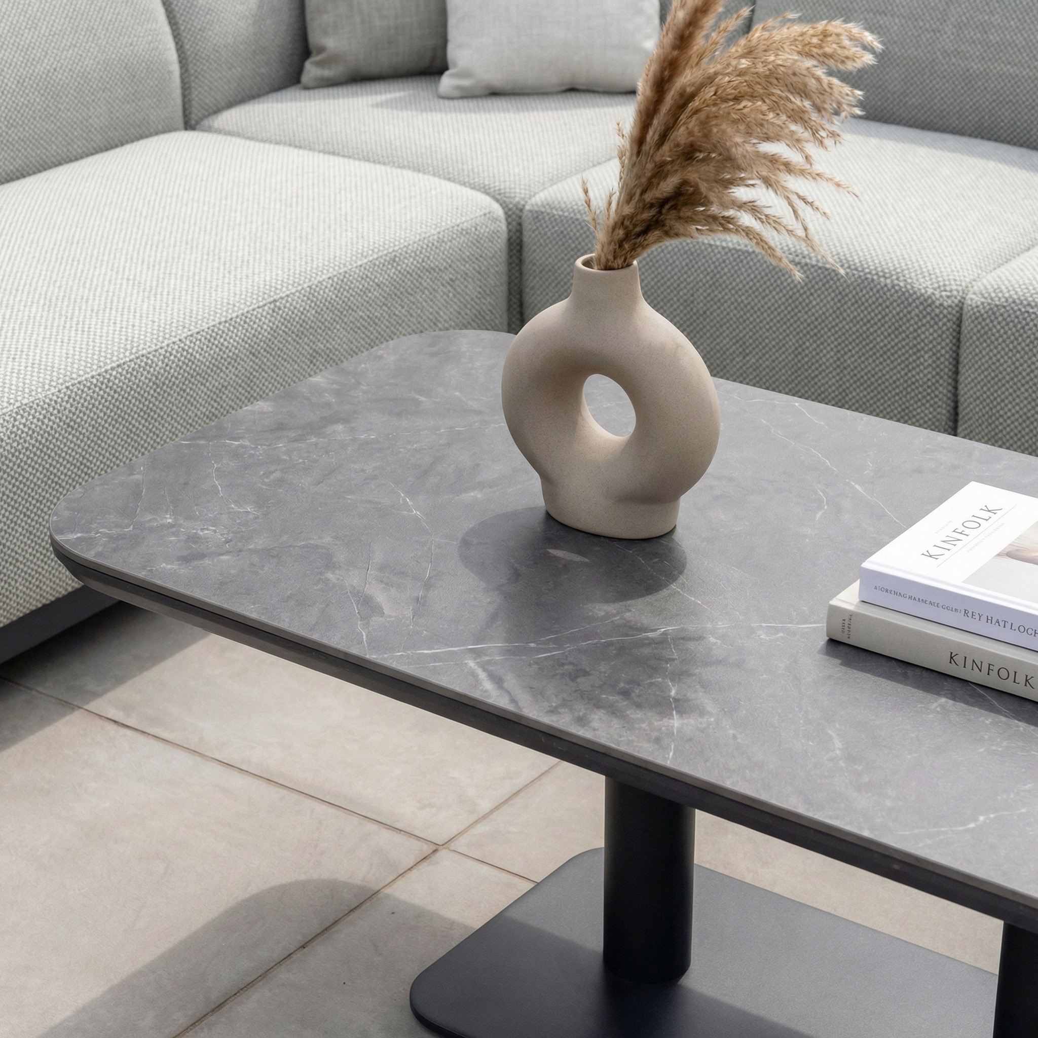 A modern coffee table with a vase of dried grasses and two books sits beside the Luna Outdoor Fabric Large Corner Sofa Set with Altura Rising Table in Oyster Grey.