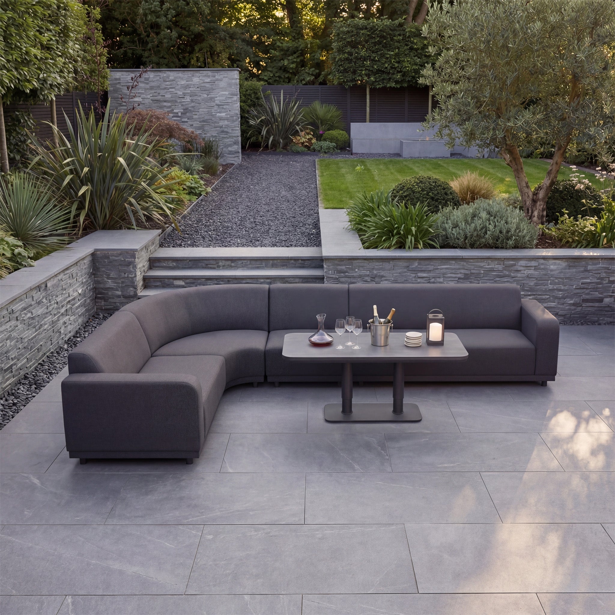 The Luna Outdoor Fabric Large Curved Corner Sofa Set with Altura Rising Table in Grey sits on a stone patio, framed by a landscaped garden. Featuring all-weather fabric, it’s perfect for stylish, durable outdoor living.