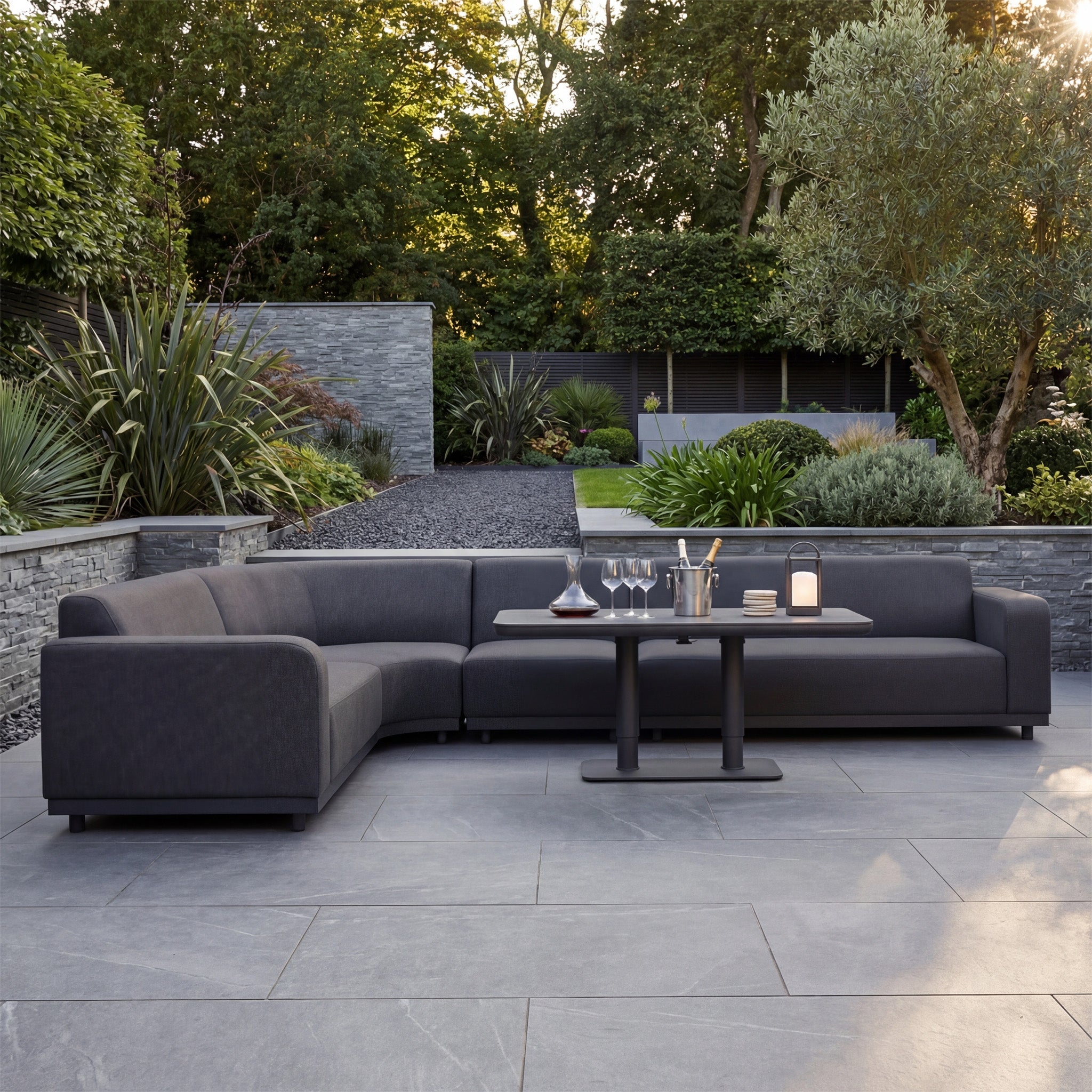 Modern patio with the Luna Outdoor Fabric Large Curved Corner Sofa Set with Altura Rising Table in Grey, drinks on the table, and a landscaped garden backdrop. Enjoy comfort and style with all-weather fabric for any season.