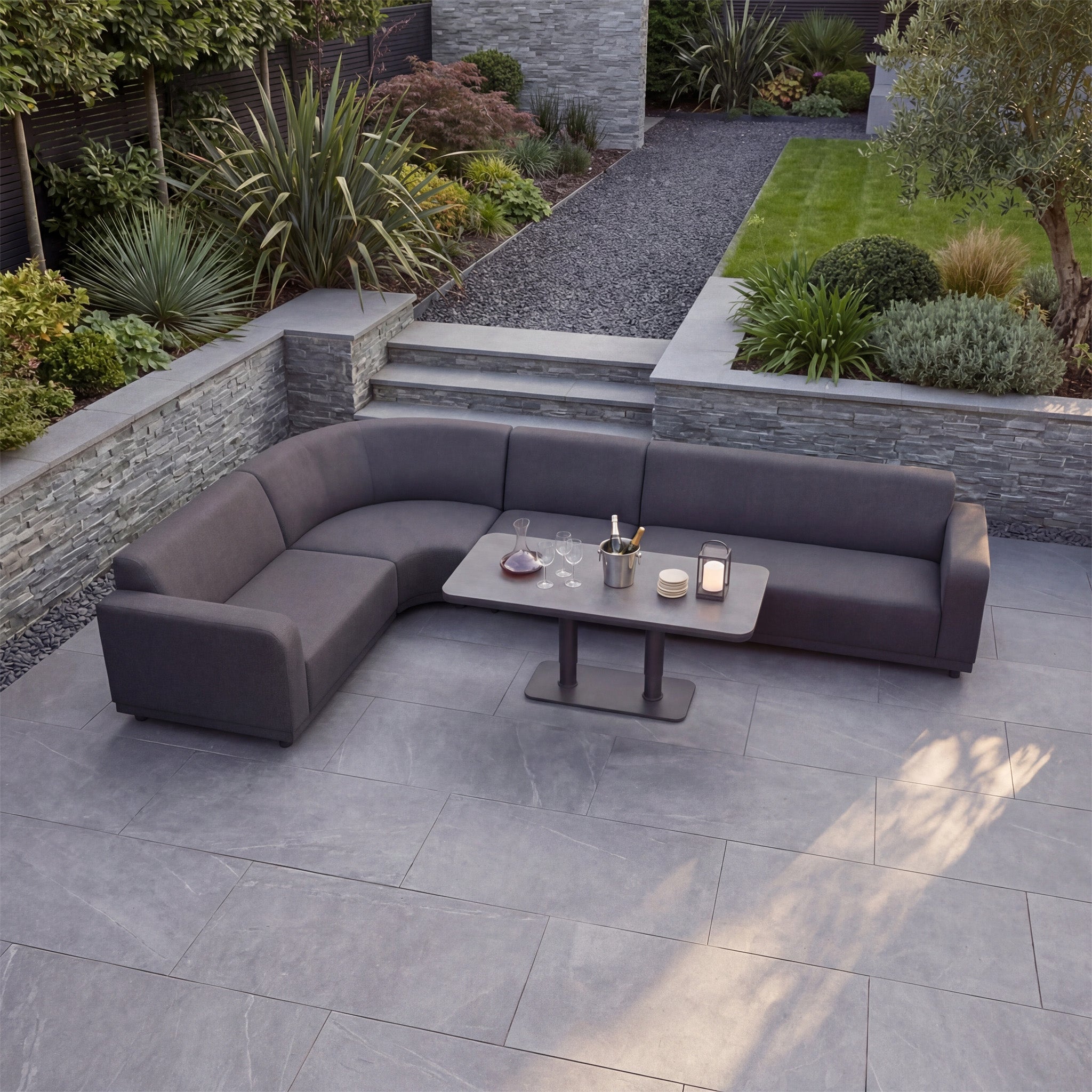 Modern outdoor patio with a grey Luna Outdoor Fabric Large Curved Corner Sofa Set and an Altura Rising Table, drinks and centerpiece on the table, surrounded by greenery.