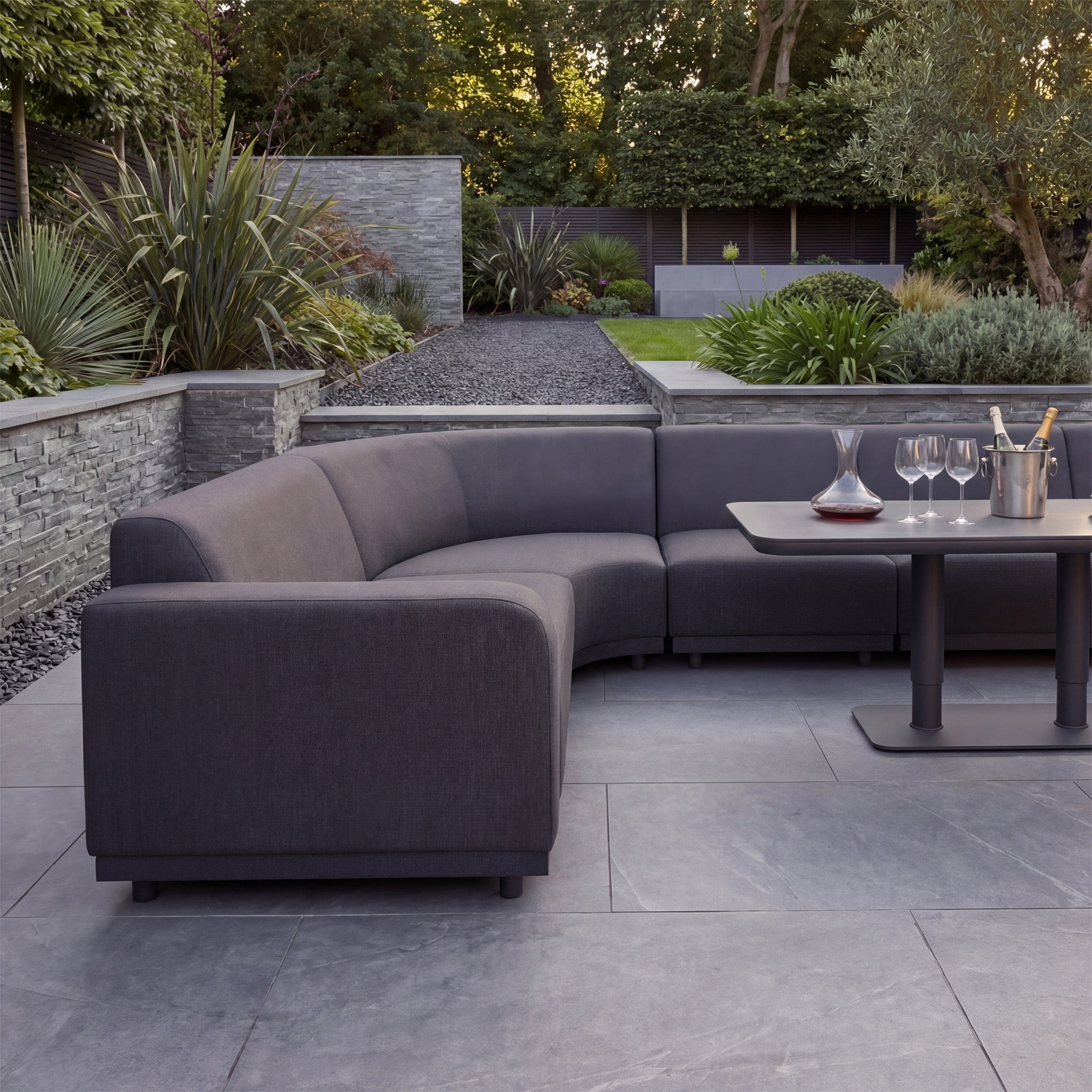 Modern patio styled with the Luna Outdoor Fabric Large Curved Corner Sofa Set and Altura Rising Table in Grey, featuring all-weather fabric, a table set with glasses, and lush greenery in the background.