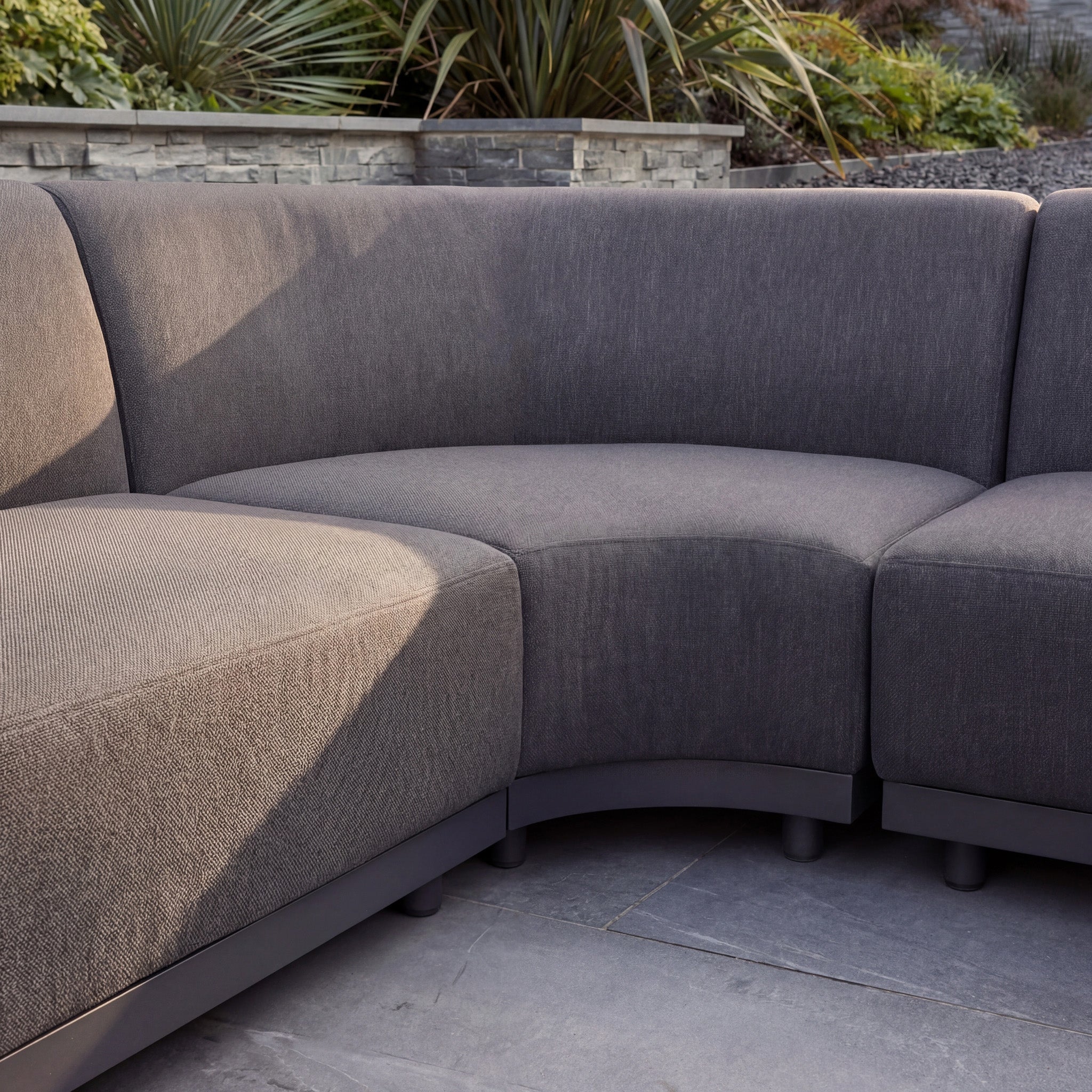 The Luna Outdoor Fabric Large Curved Corner Sofa Set with Altura Rising Table in Grey creates a stylish outdoor lounge on a stone patio, complemented by plants and a stone wall in the background.