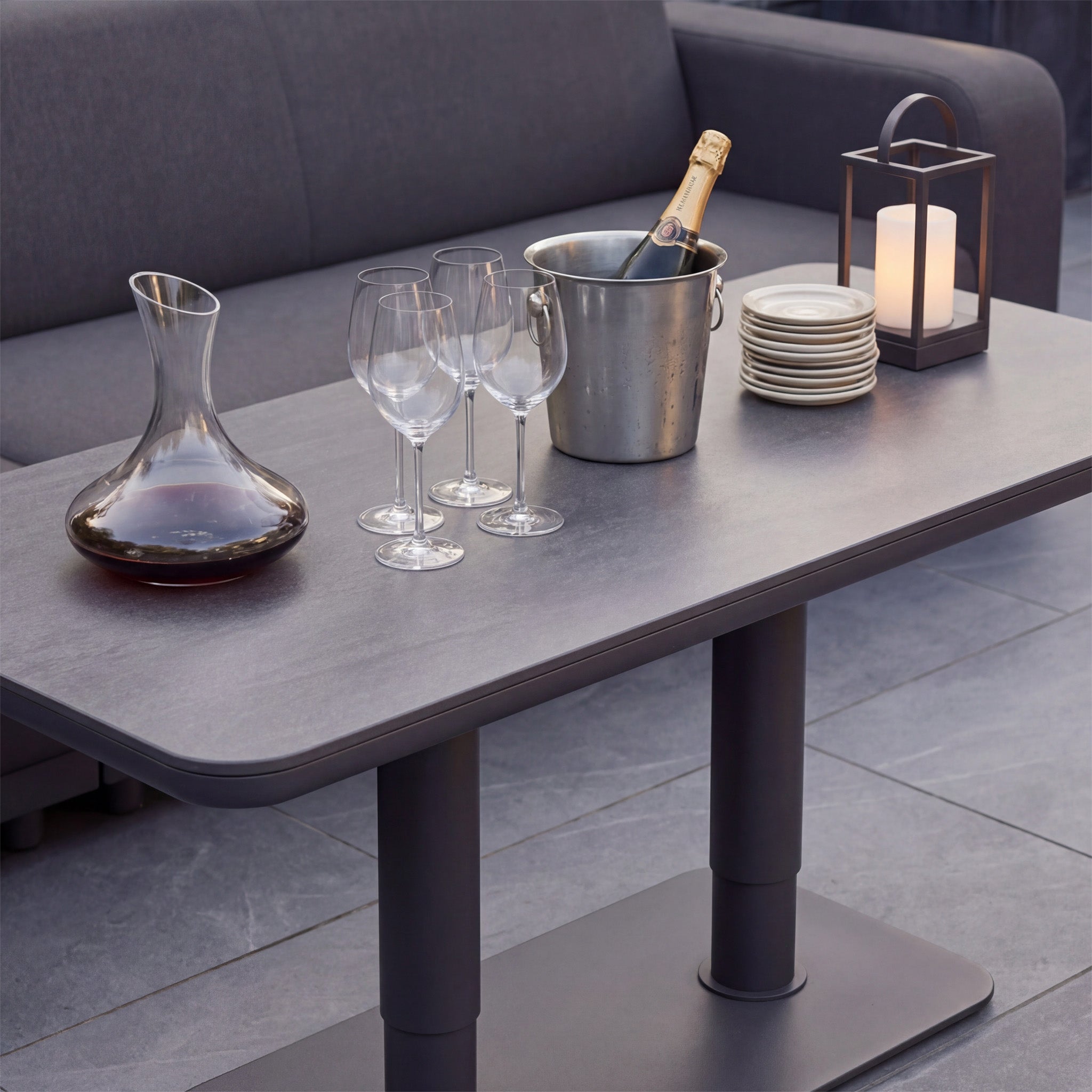 A table with wine glasses, a decanter, plates, a candle lantern, and champagne in an ice bucket sits beside the Luna Outdoor Fabric Large Curved Corner Sofa Set with Altura Rising Table in Grey.
