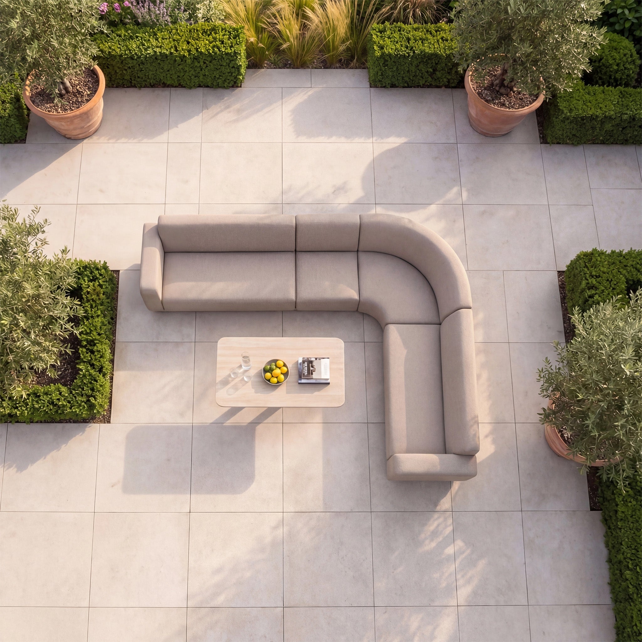 The Luna Outdoor Fabric Large Curved Corner Sofa Set with Altura Rising Table in Fawn sits on a tiled patio, surrounded by potted plants and lush hedges—an elegant all-weather garden furniture ensemble.