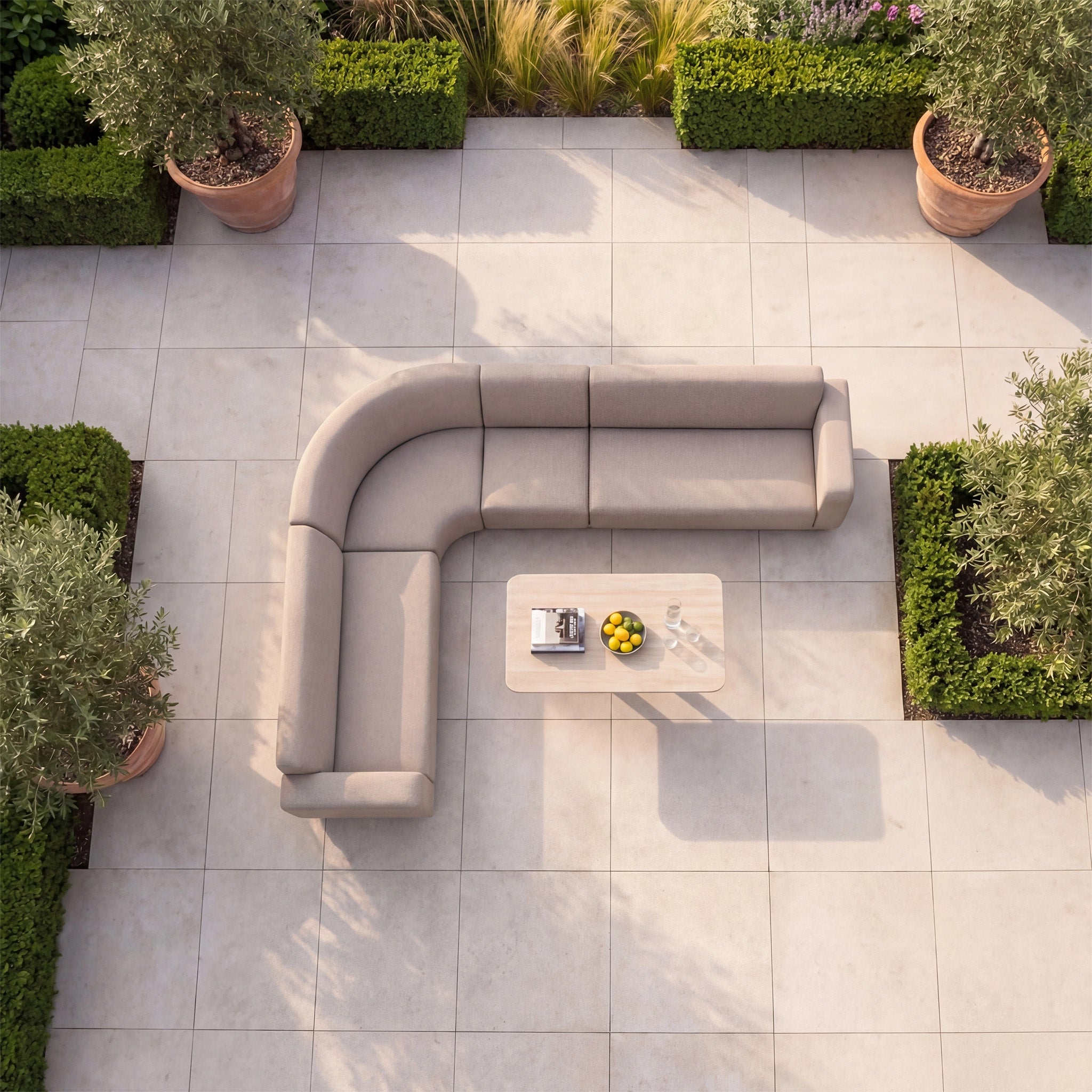The Luna Outdoor Fabric Large Curved Corner Sofa Set with Altura Rising Table in Fawn sits on a tiled patio surrounded by greenery and potted plants, viewed from above.