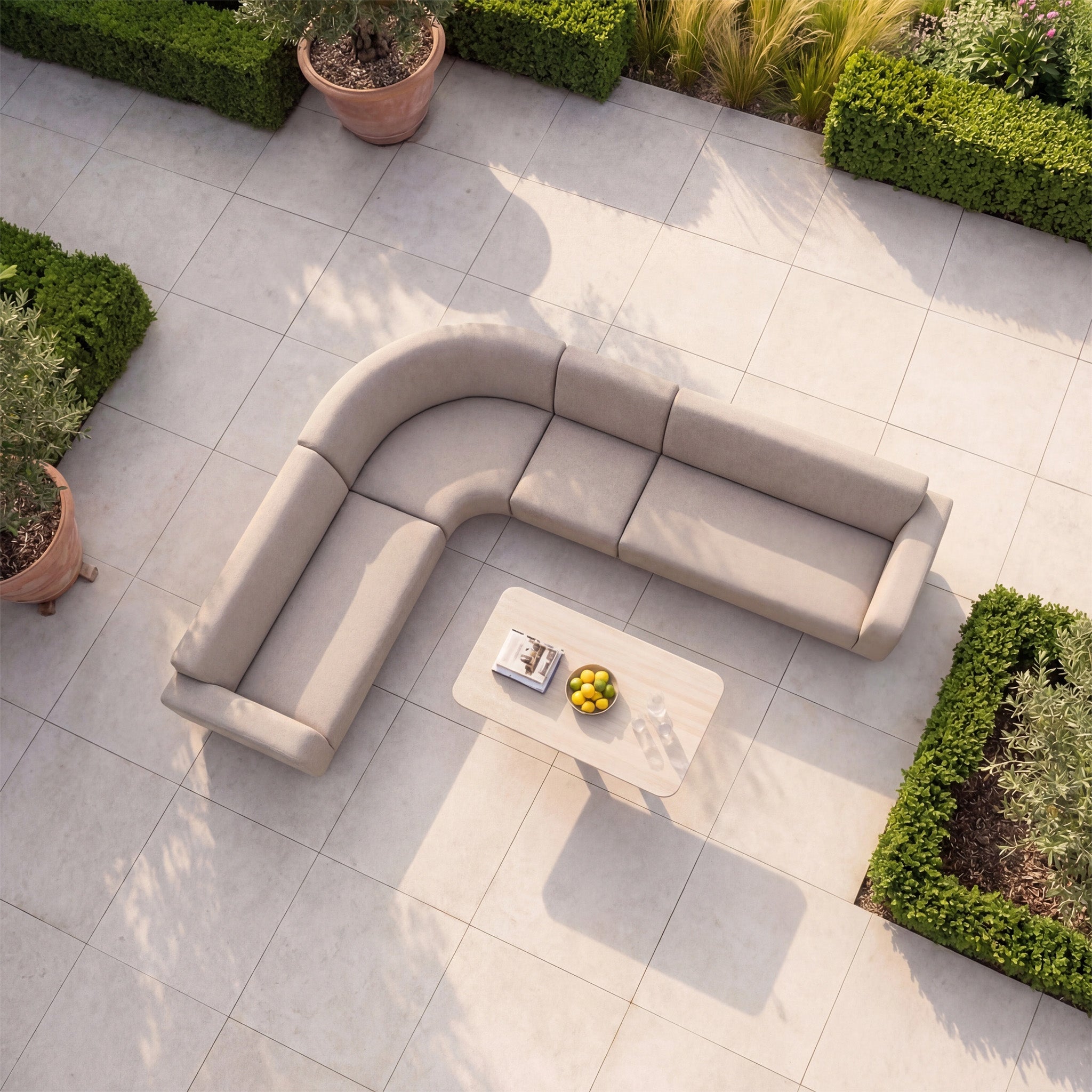 The Luna Outdoor Fabric Large Curved Corner Sofa Set with Altura Rising Table in Fawn sits on a tiled patio amid green hedges and plants—stylish, all-weather comfort for outdoor relaxation.