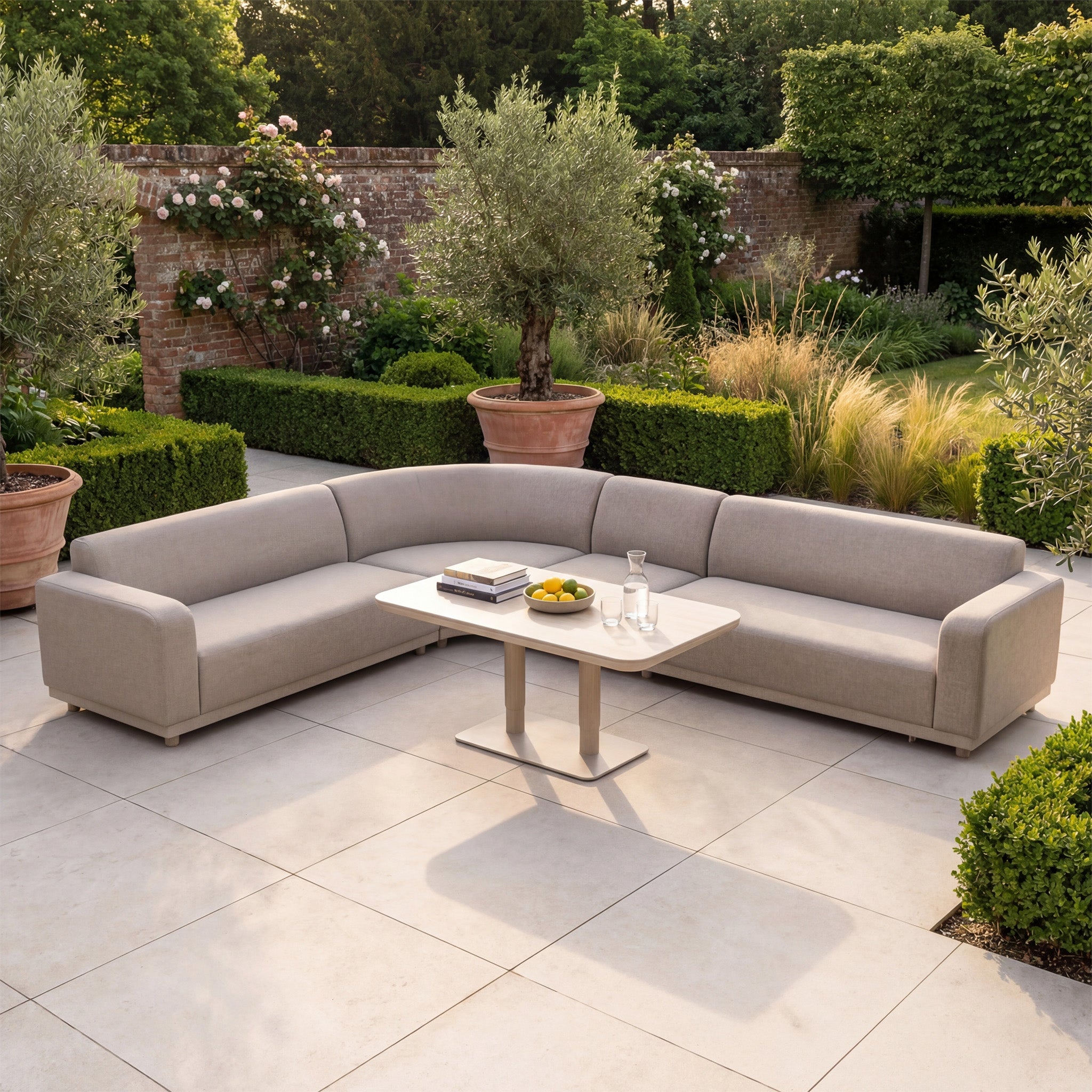 The Luna Outdoor Fabric Large Curved Corner Sofa Set with Altura Rising Table in Fawn sits on a tiled patio among lush greenery, offering a relaxing retreat with all-weather durability and comfort.