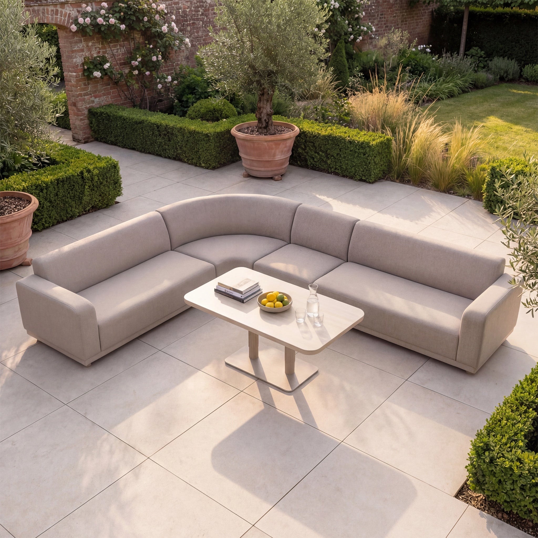 Luna Outdoor Fabric Large Curved Corner Sofa Set with Altura Rising Table in Fawn displayed in a garden with books, fruit, and water on the white table among greenery and all-weather outdoor furniture.