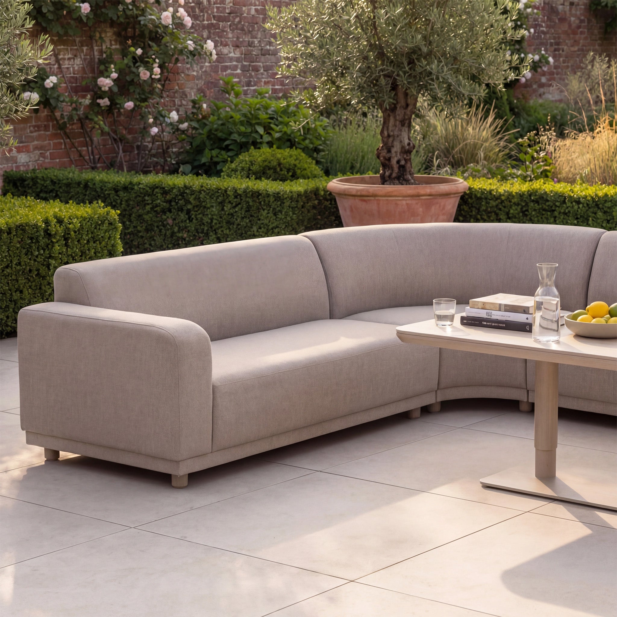 The Luna Outdoor Fabric Large Curved Corner Sofa Set with Altura Rising Table in Fawn on a tiled patio with drinks and fruit, surrounded by greenery—an ideal showcase of stylish all-weather outdoor furniture.