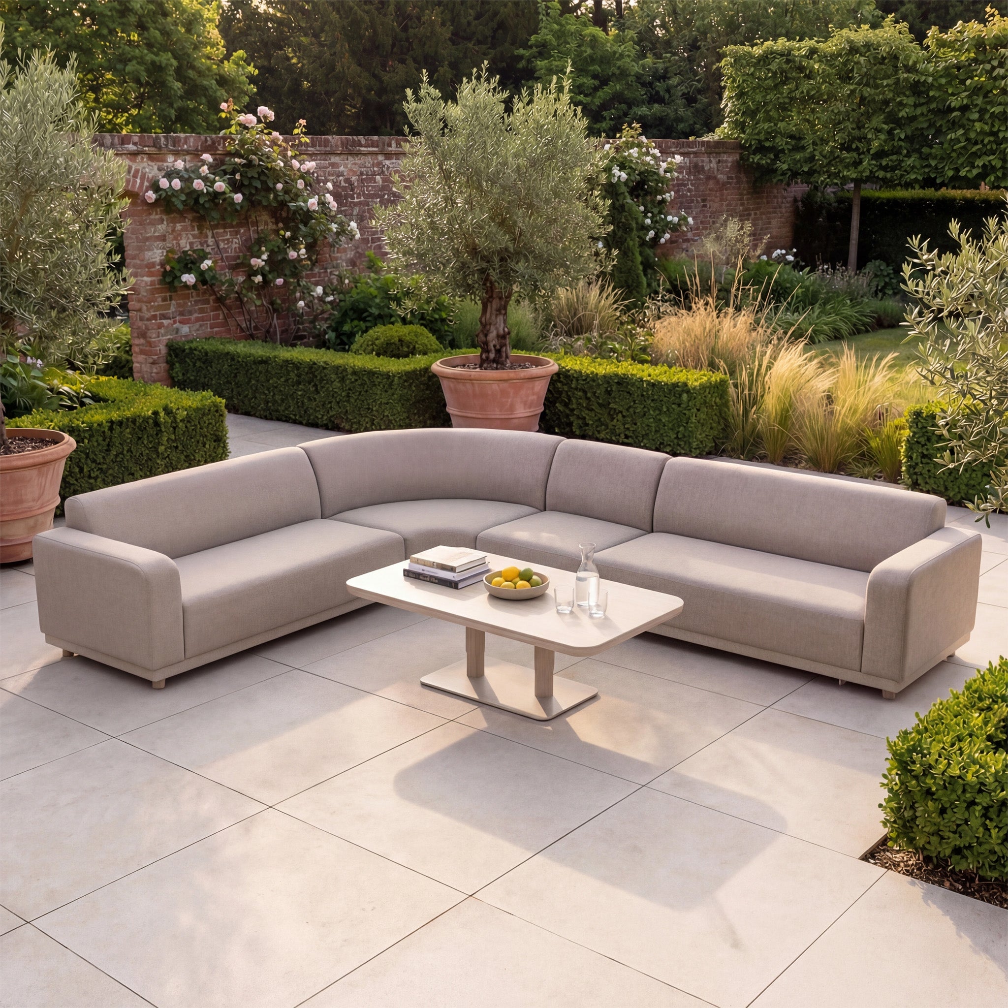 The Luna Outdoor Fabric Large Curved Corner Sofa Set with Altura Rising Table in Fawn sits on a tiled patio, encircled by lush greenery—ideal all-weather outdoor furniture for stylish, relaxing moments.