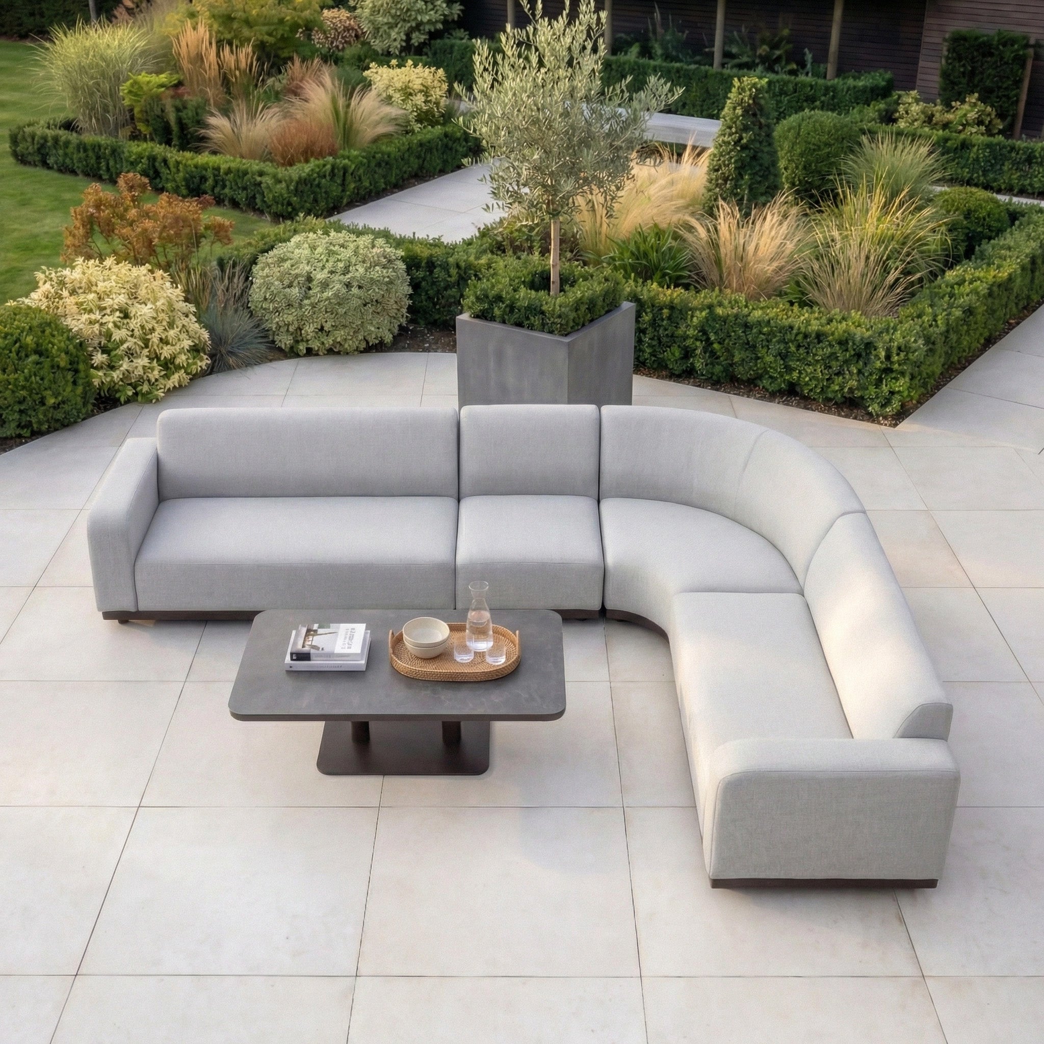 The Luna Outdoor Fabric Large Curved Corner Sofa Set with Altura Rising Table in Oyster Grey sits on a tiled patio amid lush gardens, crafted in all-weather fabric for lasting outdoor comfort.