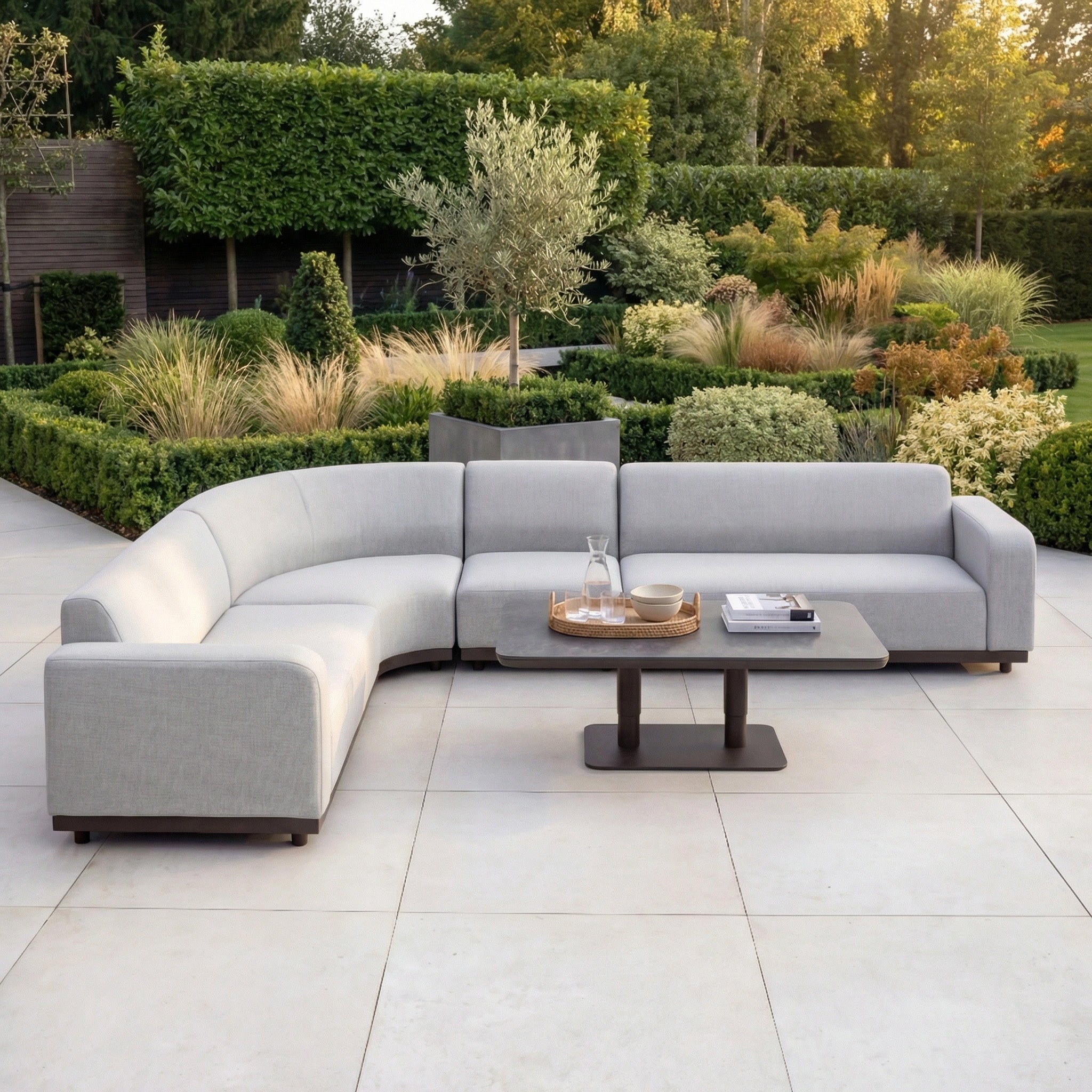 The Luna Outdoor Fabric Large Curved Corner Sofa Set with Altura Rising Table in Oyster Grey sits on a tiled patio, surrounded by a lush, green landscaped garden.