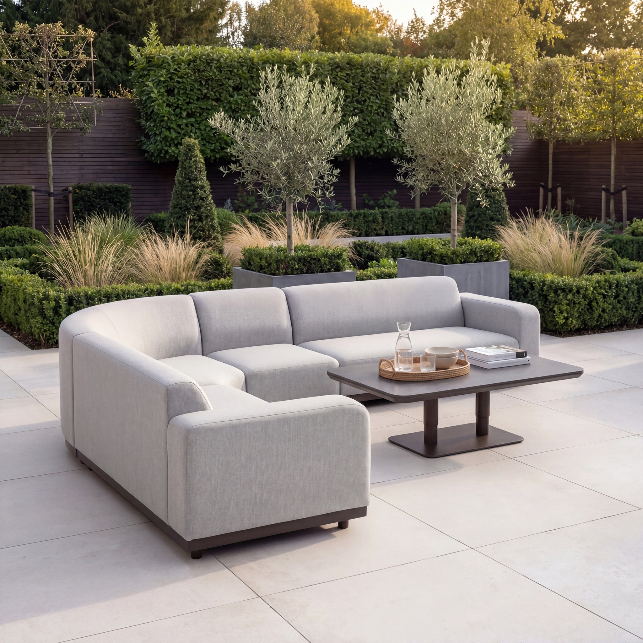 Modern outdoor patio featuring the Luna Outdoor Fabric Large Curved Corner Sofa Set with Altura Rising Table in Oyster Grey, surrounded by lush greenery and trees.