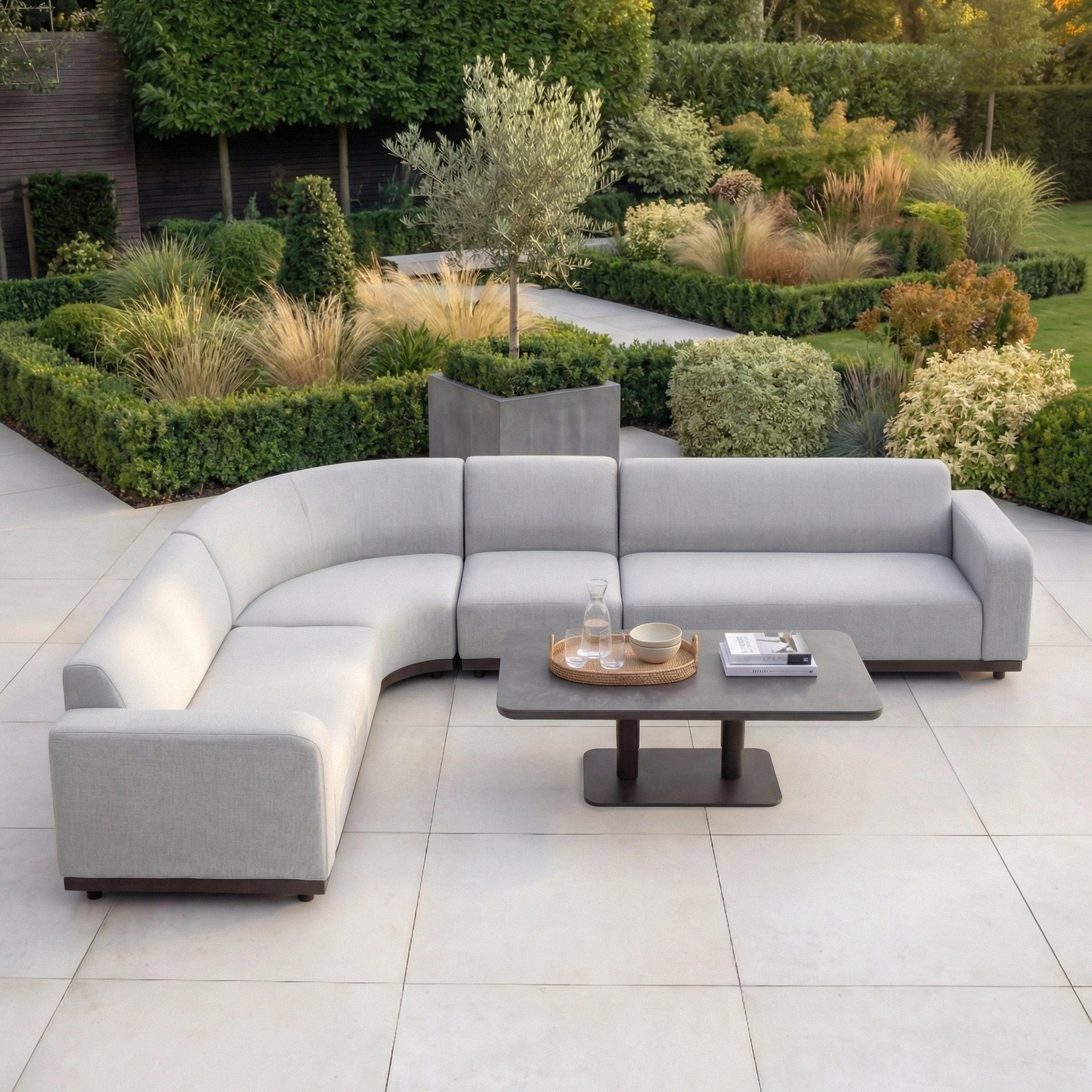 The Luna Outdoor Fabric Large Curved Corner Sofa Set with Altura Rising Table in Oyster Grey sits on a tiled patio, surrounded by landscaped greenery and bushes.