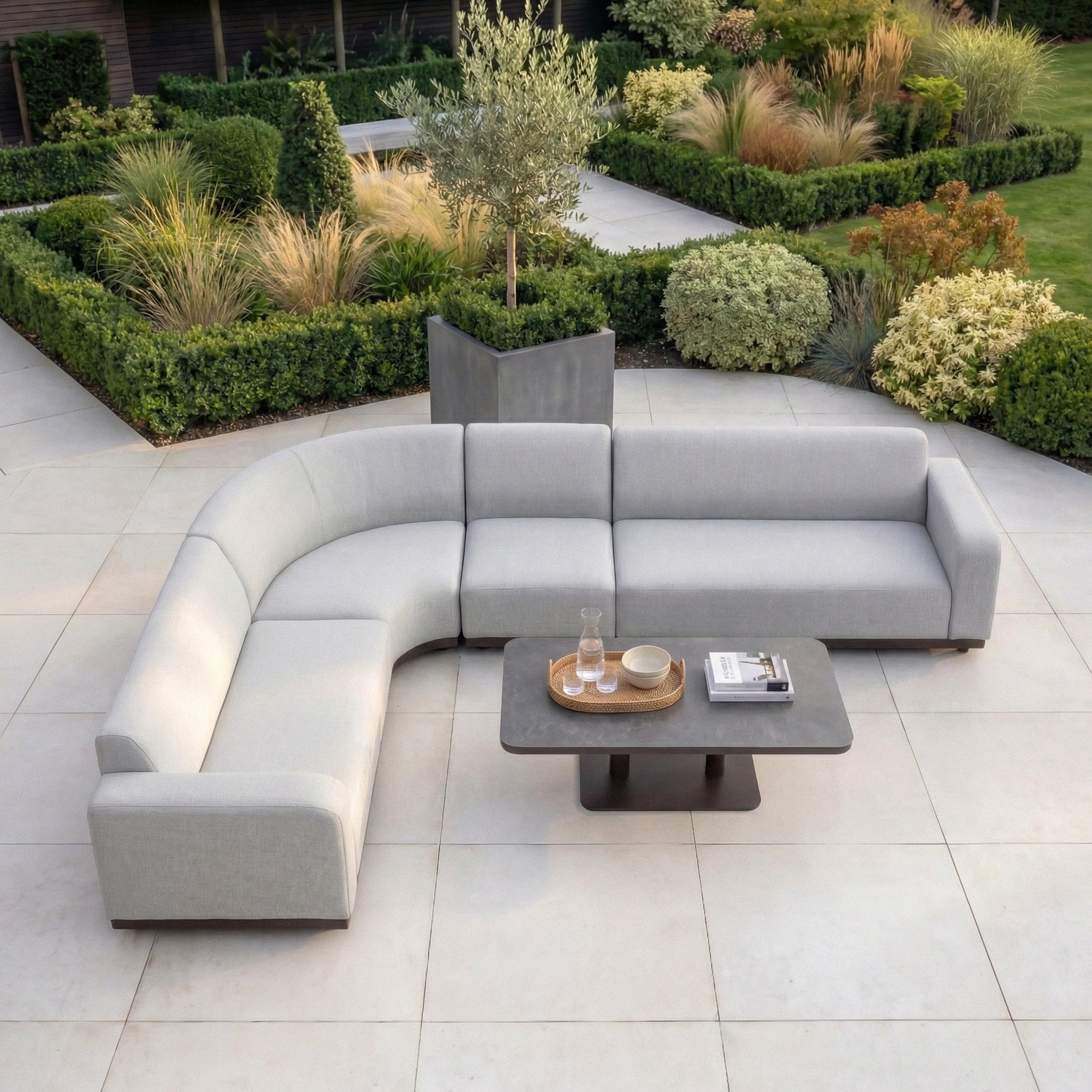 The Luna Outdoor Fabric Large Curved Corner Sofa Set with Altura Rising Table in Oyster Grey sits on a tiled patio among manicured shrubs, offering durable, all-weather comfort for year-round outdoor living.