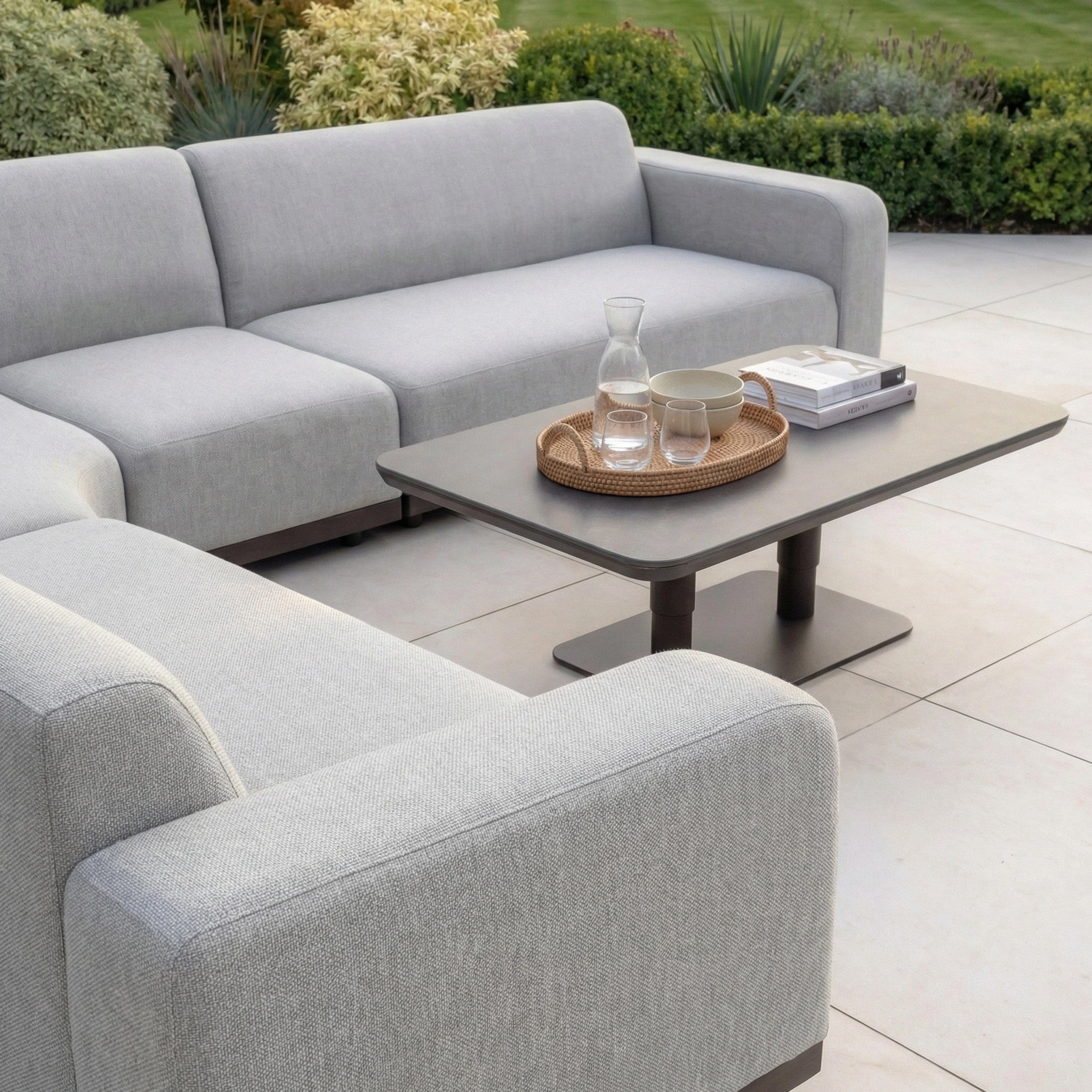 Contemporary patio with a Luna Outdoor Fabric Large Curved Corner Sofa Set in Oyster Grey and an Altura Rising Table, styled with a tray, glass pitcher, cups, and books—all made from durable all-weather fabric.