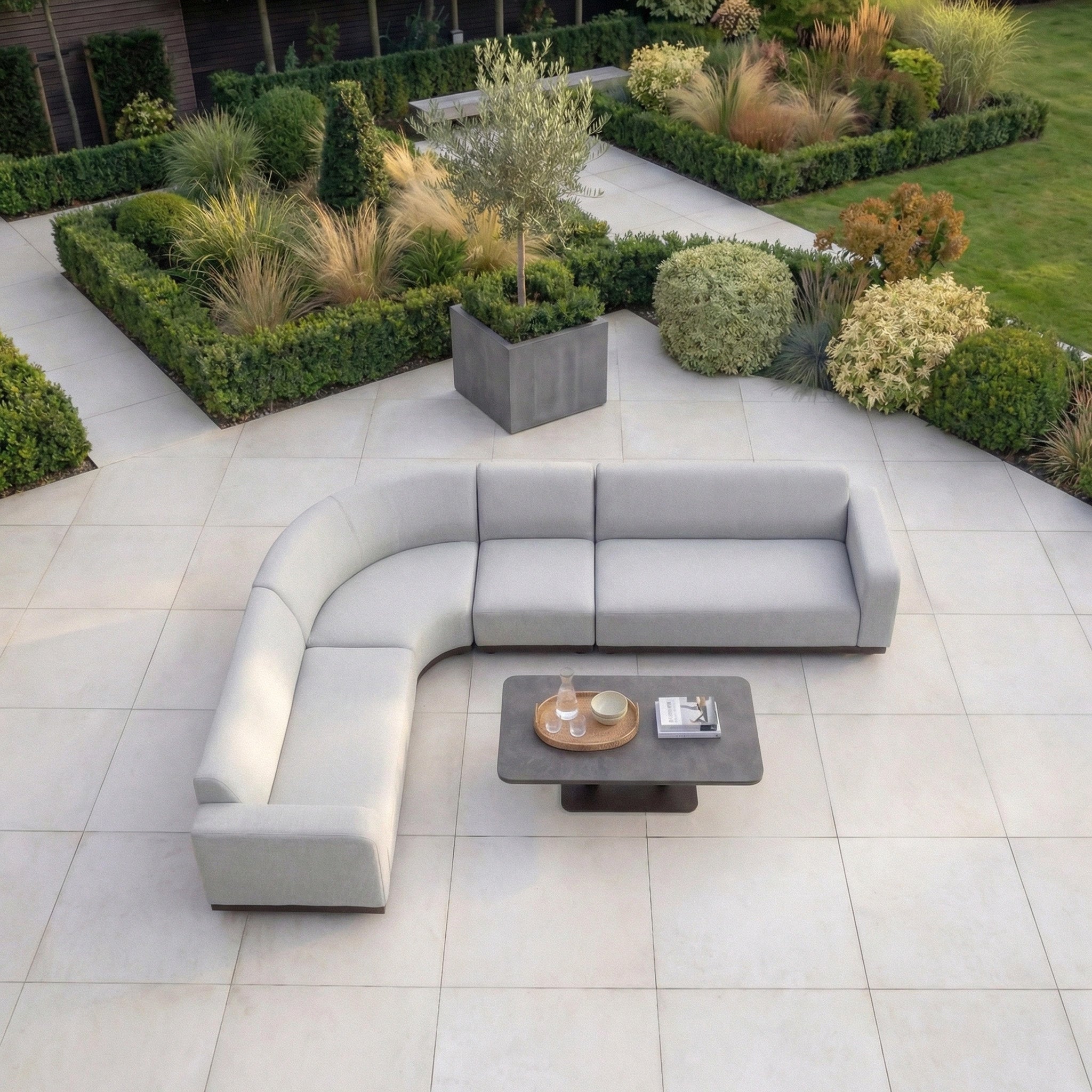 Modern outdoor patio featuring a Luna Outdoor Fabric Large Curved Corner Sofa Set with Altura Rising Table in Oyster Grey and a landscaped garden in the background.