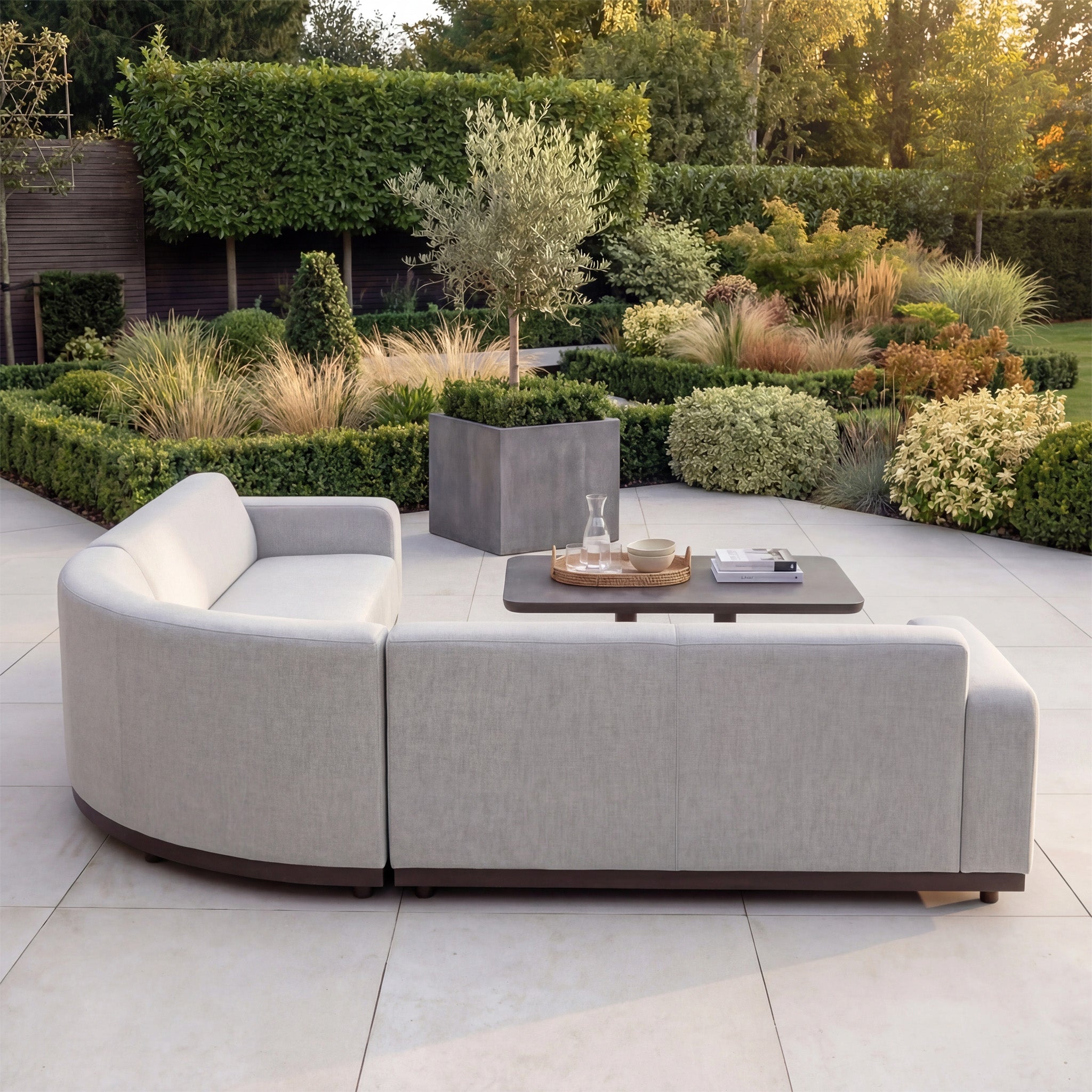 Luna Outdoor Fabric Large Curved Corner Sofa Set with Altura Rising Table in Oyster Grey on a patio, surrounded by lush landscaped garden greenery.
