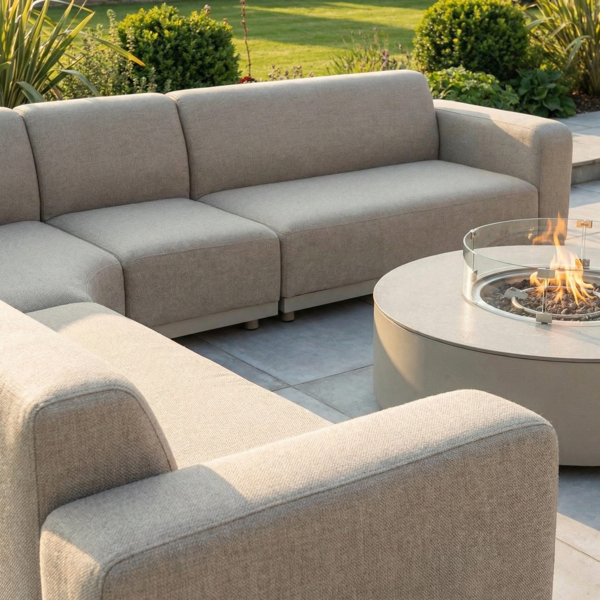 Outdoor patio featuring the Luna Outdoor Fabric Large Curved Corner Group Set with a Round Firepit Coffee Table in Fawn, surrounded by lush greenery and lawn.