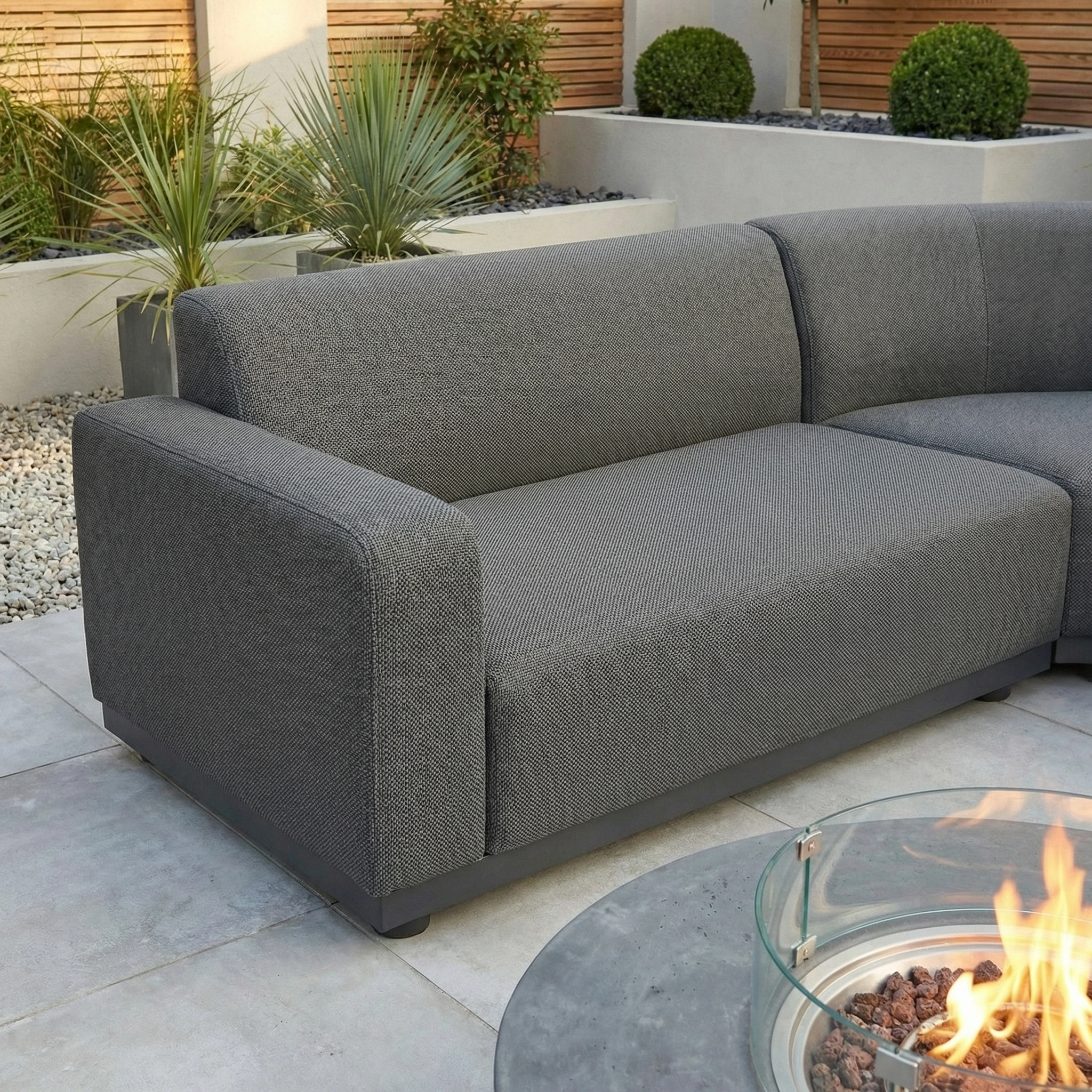 The Luna Outdoor Fabric Large Curved Corner Group Set with Round Firepit Coffee Table in Grey features quick-dry cushions on a tiled patio, surrounded by plants, with a glass fire pit coffee table in the foreground.