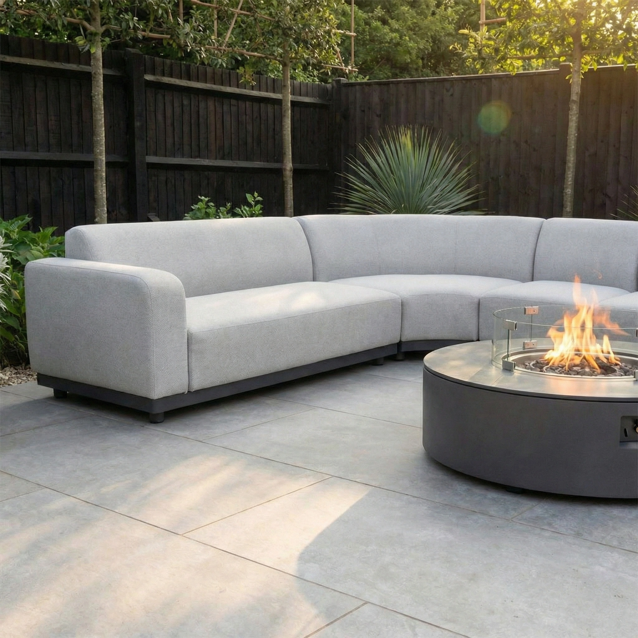 The Luna Outdoor Fabric Large Curved Corner Group Set with Round Firepit Coffee Table in Oyster Grey sits on a stone patio, surrounded by greenery.