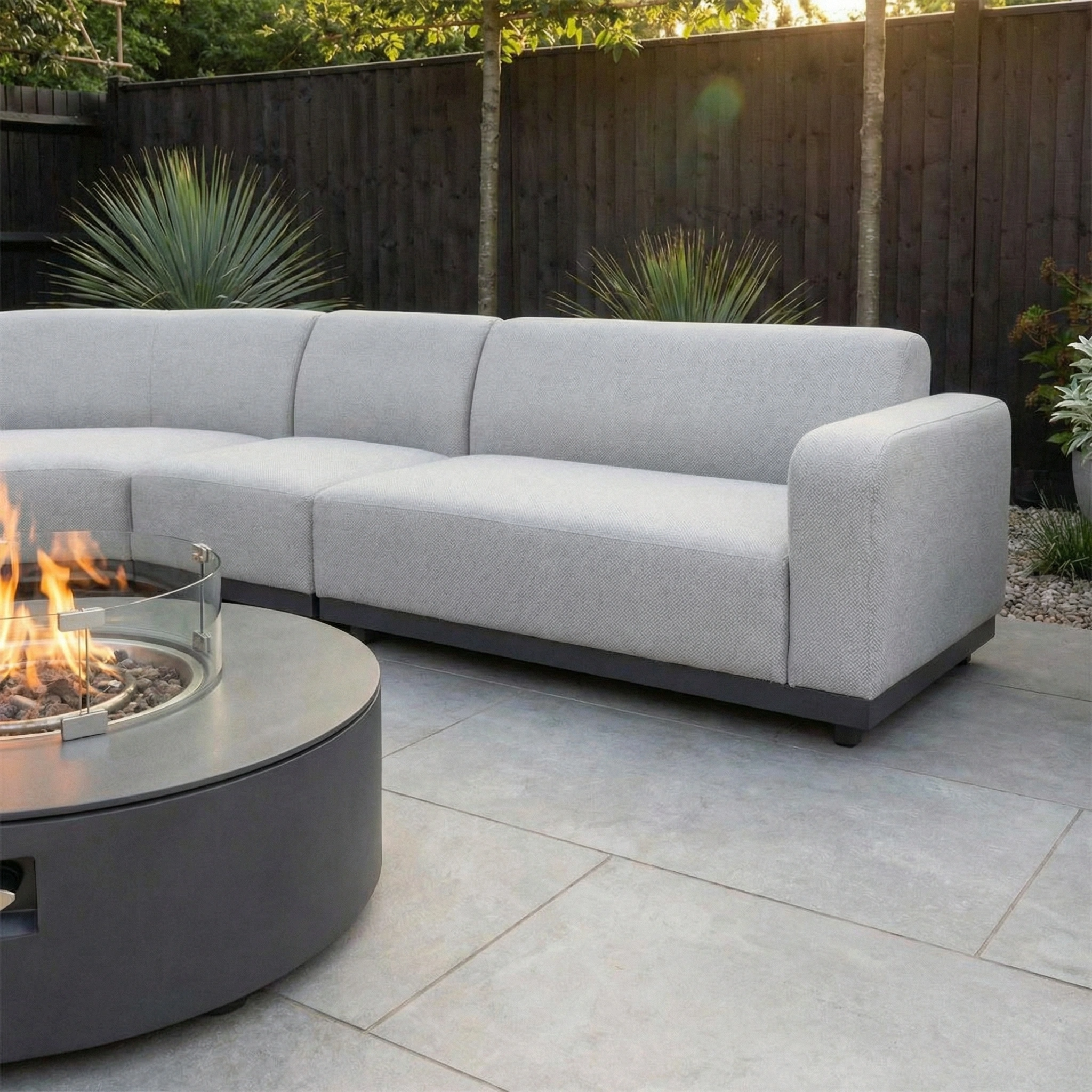 The Luna Outdoor Fabric Large Curved Corner Group Set with Round Firepit Coffee Table in Oyster Grey sits on a tiled patio, surrounded by lush plants—stylish, durable all-weather furniture designed for comfort.