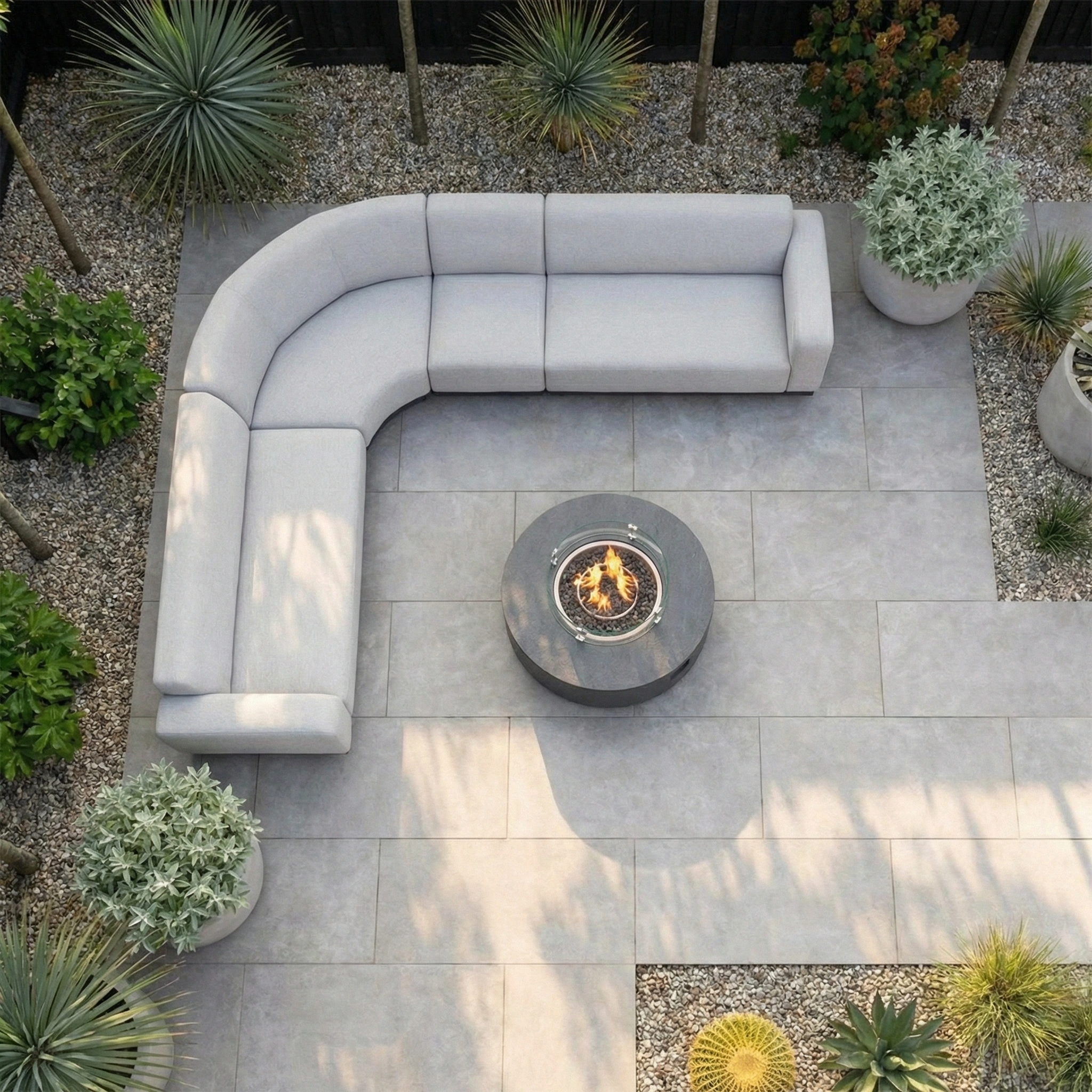 Top view of a modern patio with the Luna Outdoor Fabric Large Curved Corner Group Set and Round Firepit Coffee Table in Oyster Grey, surrounded by lush plants—perfect all-weather outdoor furniture for relaxing evenings.