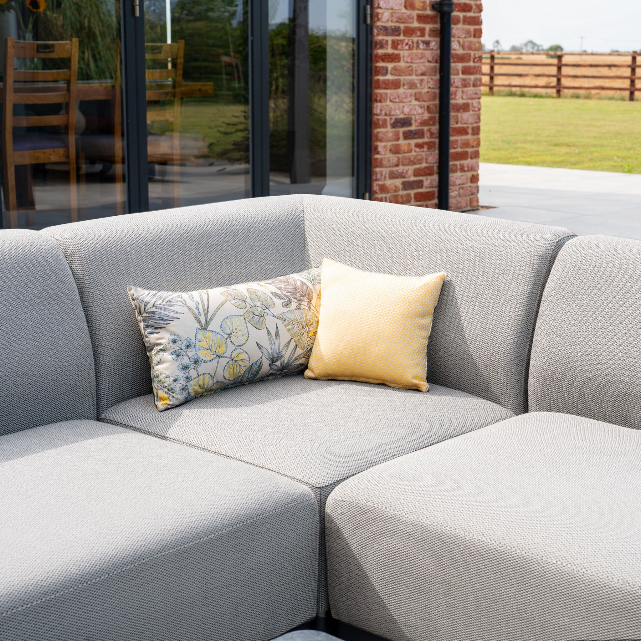 Luna Outdoor Fabric Large Corner Group Set with Coffee Table in Oyster Grey, featuring two decorative pillows—an elegant all-weather garden sofa placed near glass doors and a brick wall.