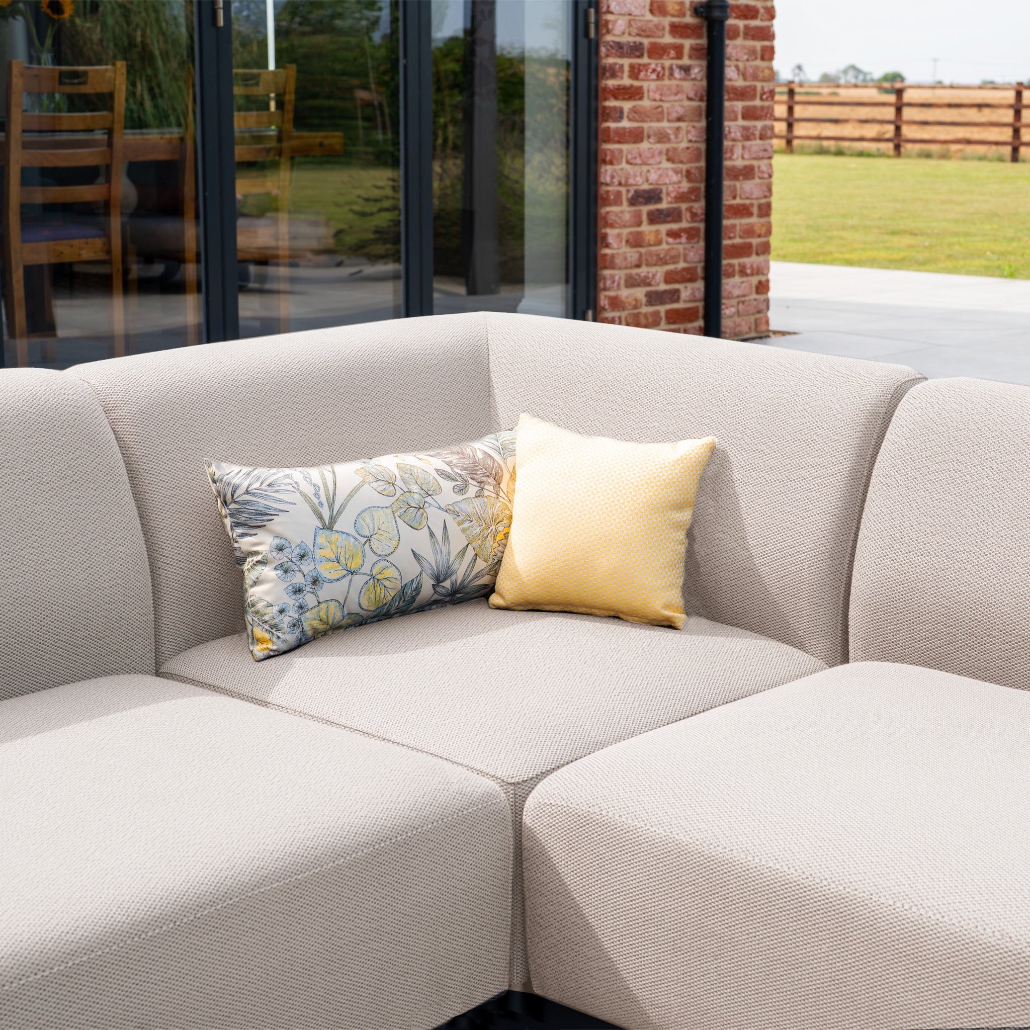 The Luna Outdoor Fabric Large Corner Group Set with Coffee Table in Fawn, featuring all-weather comfort and two decorative pillows, is displayed before glass doors and a brick wall.