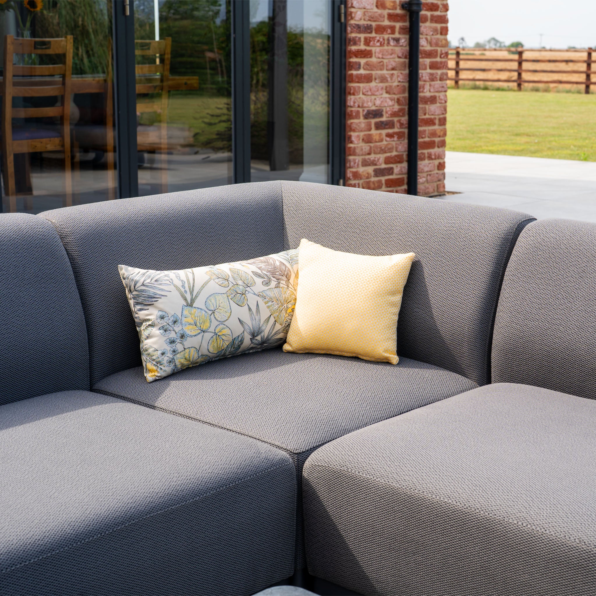 The Luna Outdoor Fabric Corner Sofa Set with Altura Rising Table in Grey, featuring stain-resistant fabric and two decorative pillows, is ideal patio furniture for relaxing by glass doors and a brick wall.