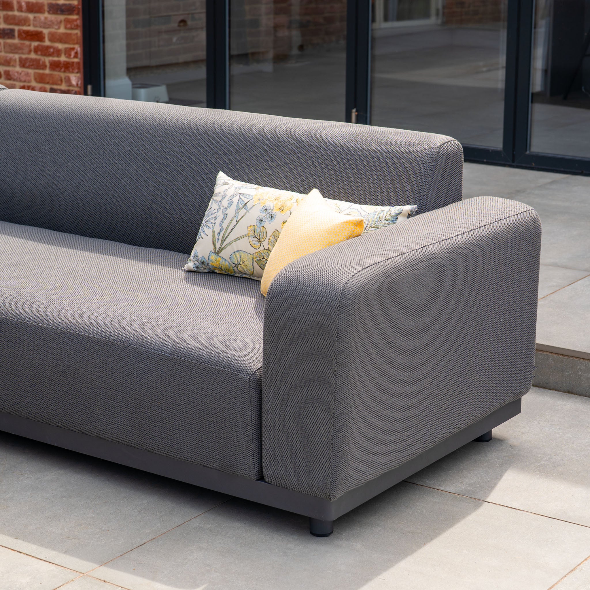 The Luna Outdoor Fabric Corner Sofa Set with Altura Rising Table in Grey, featuring stain-resistant fabric and decorated with yellow and floral pillows, is placed on the patio near the glass door.